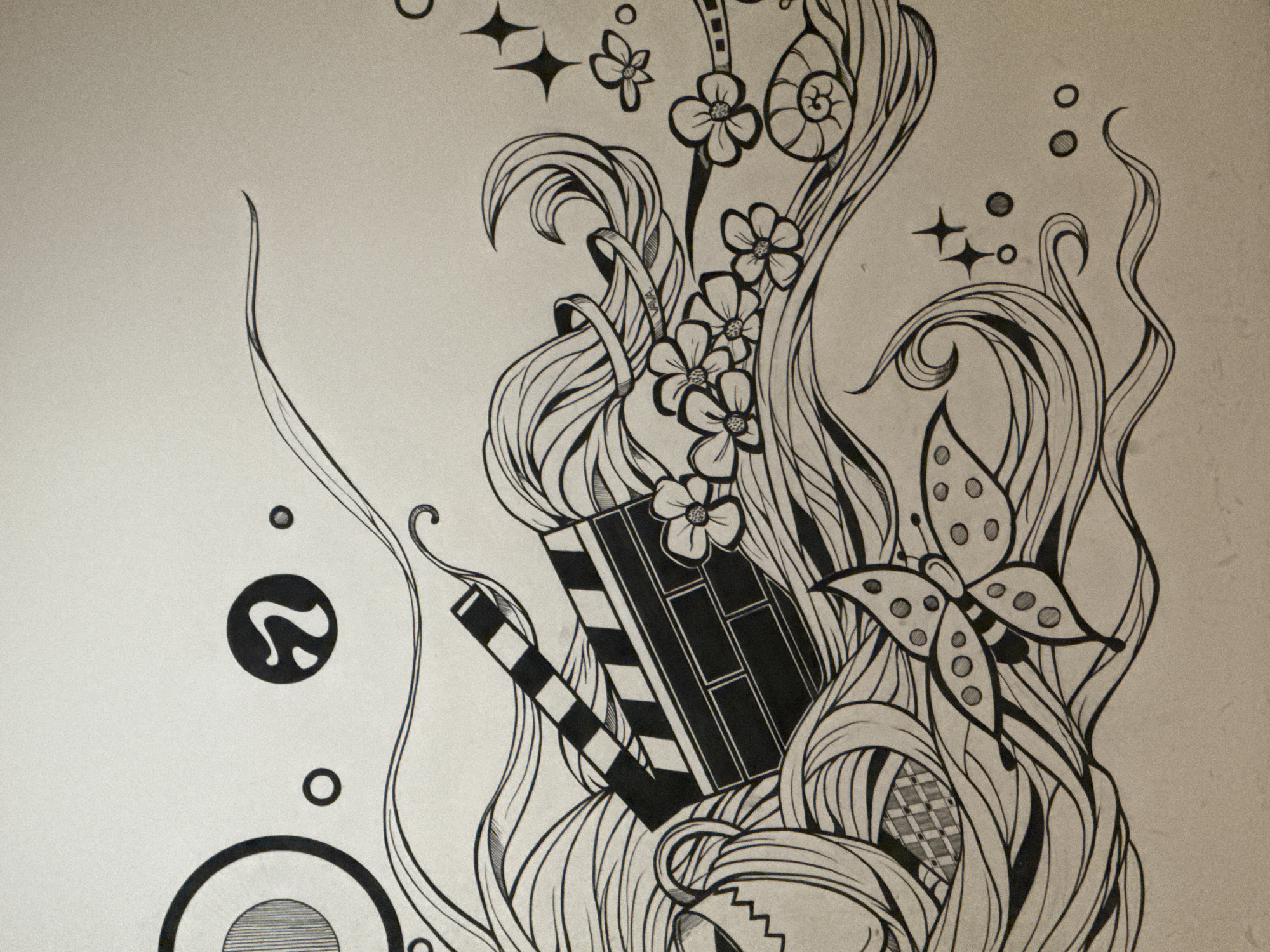 An ink drawing on a tan background depicts a whimsical and abstract scene. The composition is filled with flowing lines that create a sense of movement, resembling water or wind. Prominently featured is a clapperboard, a symbol often associated with filmmaking, angled diagonally across the drawing. To the left of the clapperboard, a teacup with a zig-zag pattern is partially visible. Scattered throughout the scene are stylized flowers, a snail shell, a butterfly with dotted wings, and several circular and star-shaped elements. Towards the top left, a large circle with radial lines encloses a smaller, similarly lined circle. A distinct black symbol resembling a stylized "2" or a curling ribbon is also present in the upper portion of the image. The drawing is executed in black ink with varying line weights, creating depth and texture.