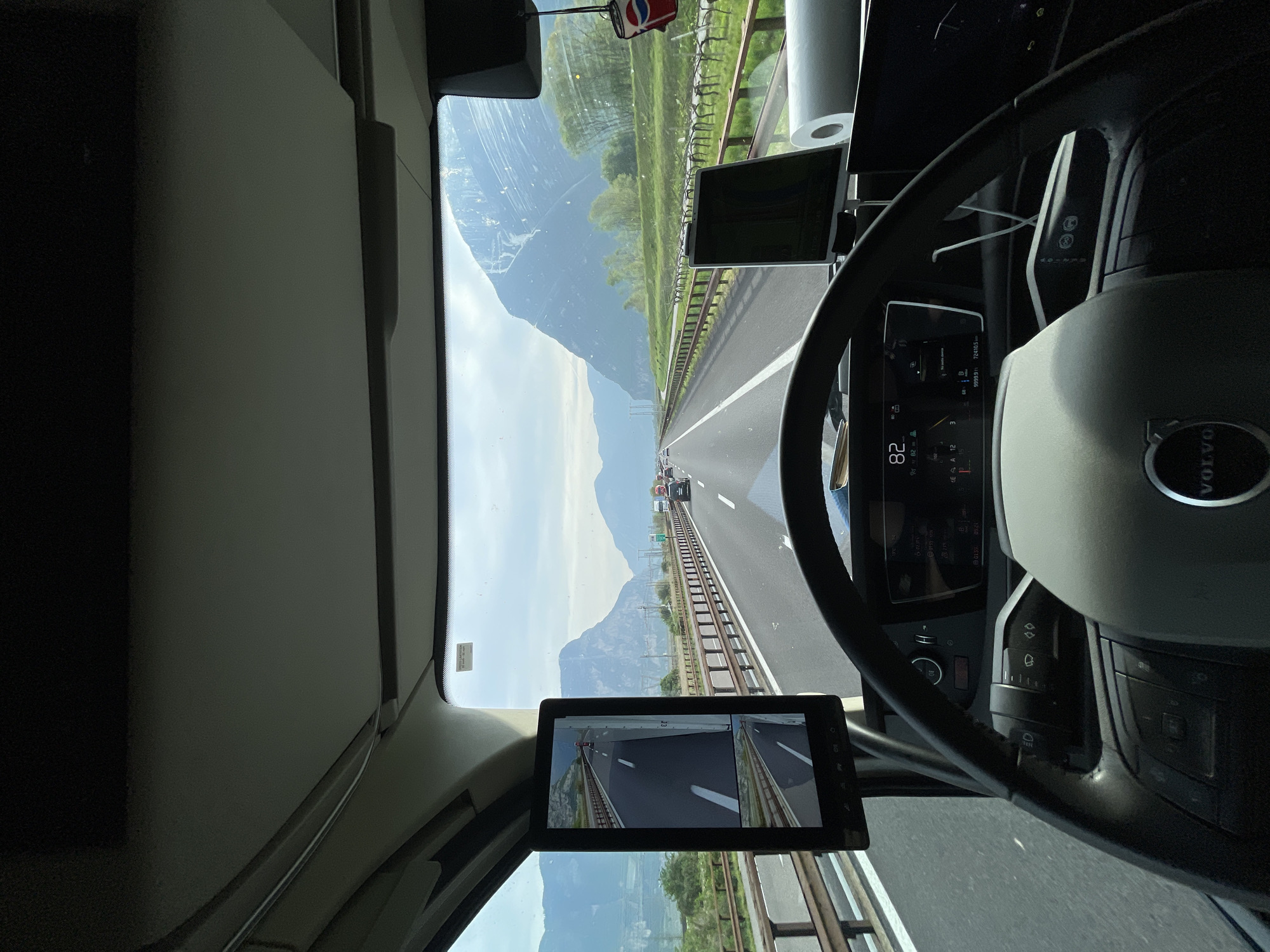 The image is a view from the driver's seat of a Volvo truck traveling on a highway near Mezzocorona, Italy. The speedometer reads 82. Mountains are visible in the distance. A monitor in the cab shows rear views. A roll of paper towels is hanging from the cab's ceiling.