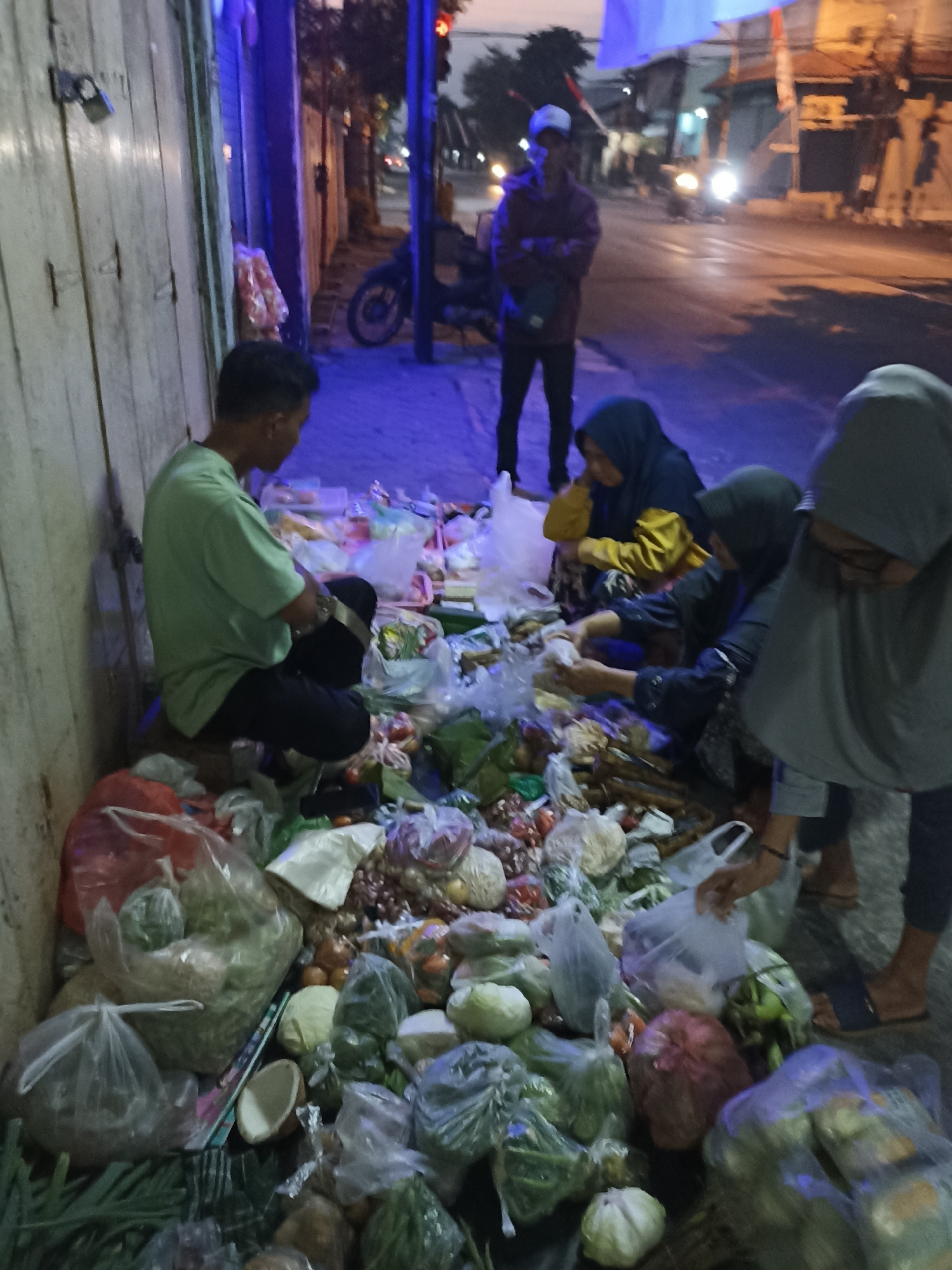 A group of people are gathered around a large assortment of vegetables and fruits displayed on a sidewalk at night, likely a street market in Lumajang, Indonesia. A man wearing a green shirt and dark pants sits on a wooden stool, appearing to be organizing or preparing the produce. To his right, three individuals are interacting with the vegetables. One person in a blue hijab and yellow shirt is looking down, while another in a dark hijab and jacket is handling items. A third person wearing a grey hijab is partially visible, also engaged with the produce. The street is illuminated by artificial lights, casting a bluish hue on the scene. A motorcycle is parked on the sidewalk behind the individuals. The ground is covered with plastic bags and various types of vegetables such as onions, cabbage, and leafy greens. The overall scene suggests a commercial transaction or preparation for sales on a busy urban street.