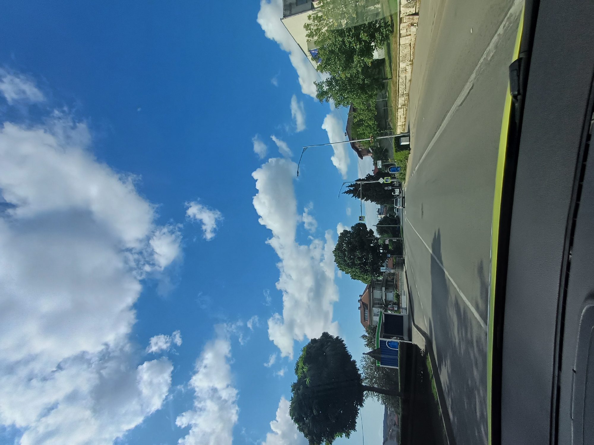 This image depicts a street scene in Aksakovo, Bulgaria, likely during the daytime given the bright sunlight and clear blue sky with scattered white clouds. The perspective is from inside a vehicle, indicated by the dashboard on the right side of the frame and the windshield through which the scene is viewed.
The street appears to be a two-lane road with a white line marking the center. Along the right side of the road, there are buildings and trees. One building has a light-colored facade and is partially obscured by a leafy green tree. Further down the street, more residential buildings with reddish-brown roofs are visible, interspersed with trees. There are also streetlights and traffic signals, suggesting this is a developed area. On the left side of the road, a large, dark green tree dominates the foreground. Further beyond, more trees and buildings are visible, and the sky is a vibrant blue with fluffy white clouds.
Some visible text includes street signs. One sign appears to be a pedestrian crossing sign. Another sign has a yellow background with a black symbol, possibly indicating a warning or information. A blue and green structure, possibly a bus stop shelter, is also present. The overall impression is one of a pleasant, sunny day in a town or suburban setting. No people are clearly visible.