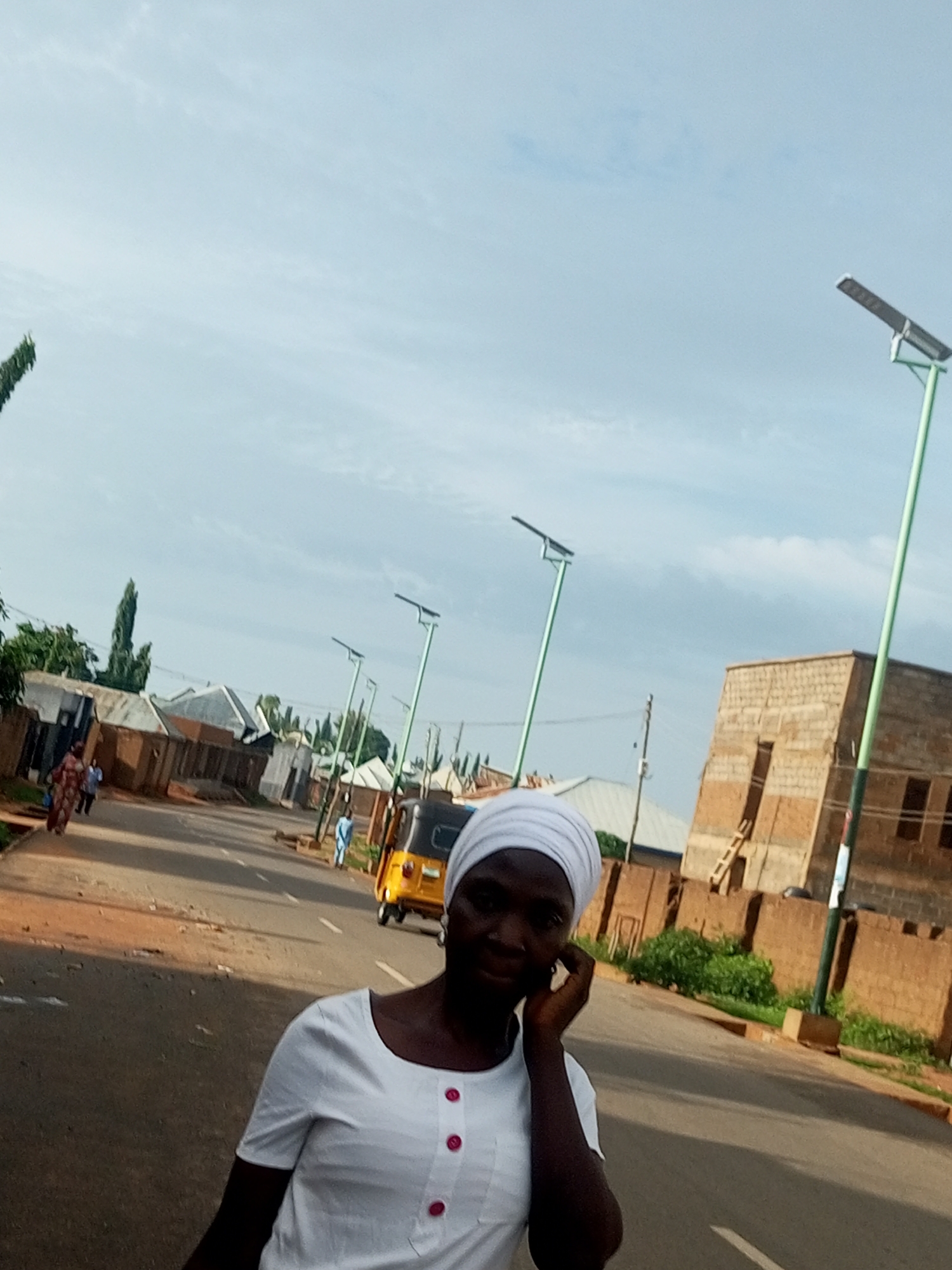 A woman wearing a white headscarf and a white shirt with pink buttons is in the foreground, facing forward and looking towards the camera. She has her hand on her right ear. Behind her, a street runs through a residential area. On the left side of the street are several low-rise buildings with terracotta-colored roofs. Several solar-powered streetlights are visible along the road. A yellow autorickshaw is driving away from the camera on the right side of the road. Further down the street, more buildings and vegetation are visible. The sky is overcast. The location is identified as Tempure, Nigeria.
