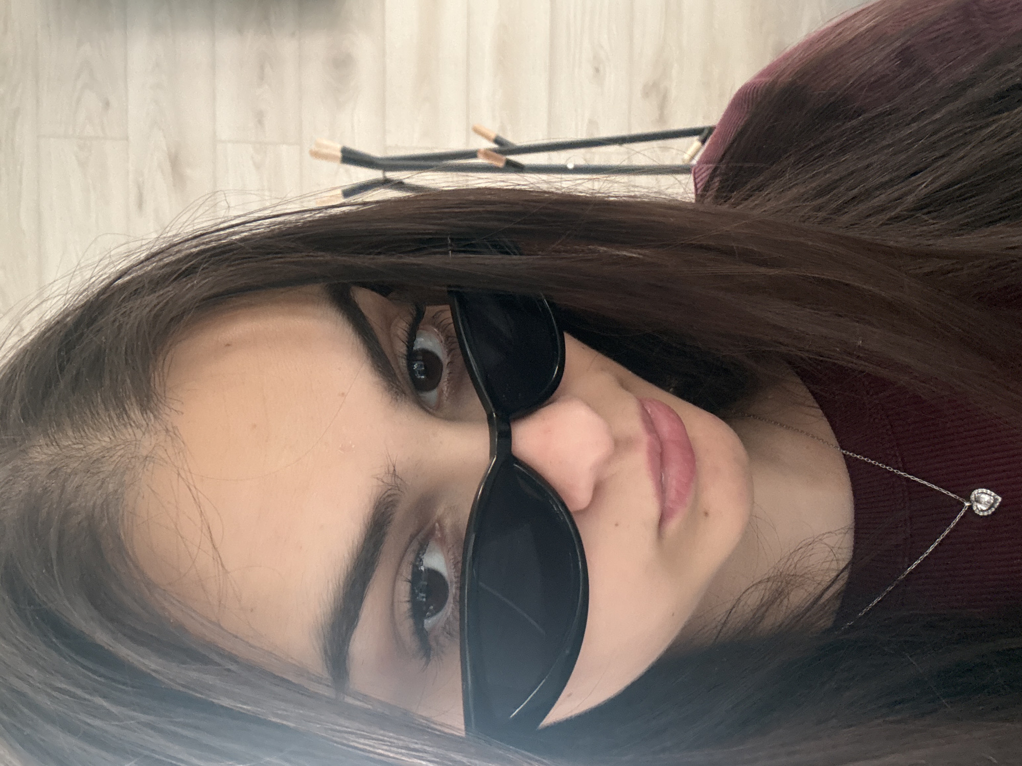 A close-up, rotated portrait shows a young woman with dark brown hair and dark eyes wearing black sunglasses. She has fair skin and pink lips. A silver necklace with a heart-shaped pendant is visible on her neck. In the background, out of focus, are wooden flooring and what appear to be makeup brushes or similar items. There is no information available to place the subject in Blagoevgrad, Bulgaria, or any other location.