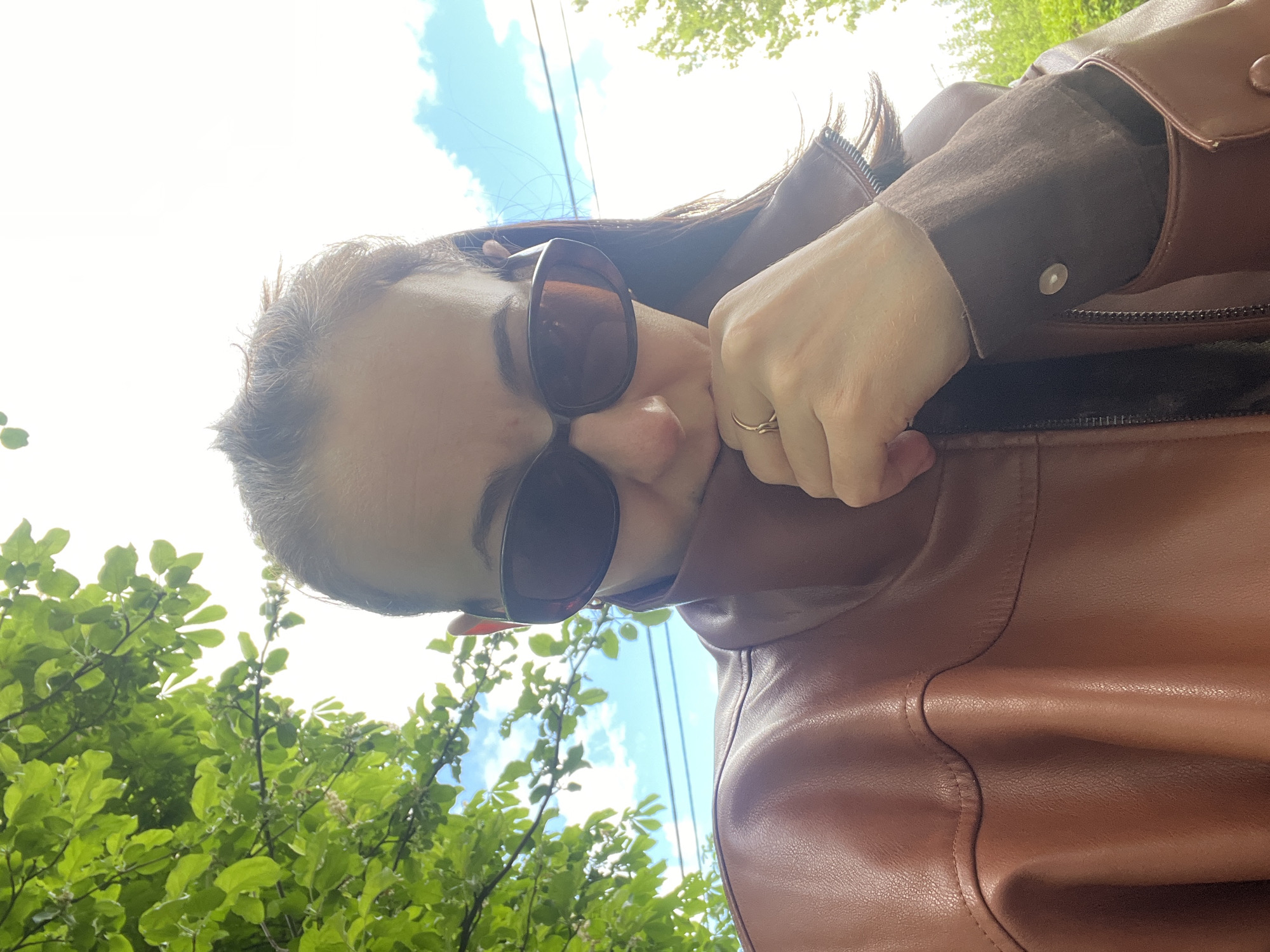 A woman wearing sunglasses and a brown leather jacket is taking a selfie from a low angle. Behind her are green leafy trees and a blue sky with white clouds. The location appears to be outdoors in a city of unknown, unknown.