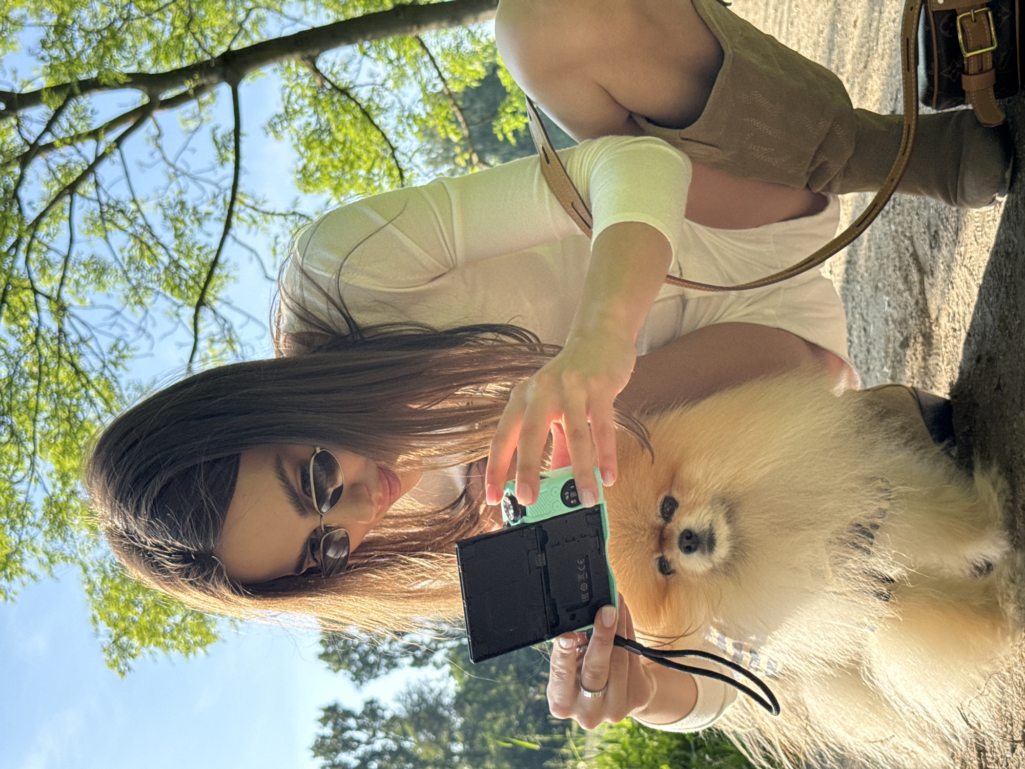 A young woman is crouching outdoors on a sunny day, holding a digital camera and appearing to take a selfie or photo of herself and a Pomeranian dog. She is wearing sunglasses, a long-sleeved white shirt, white shorts, and tall beige boots. A small purse is near her feet. The setting appears to be a park or natural area, with green foliage and trees overhead against a clear blue sky. The location is Unknown, Unknown.