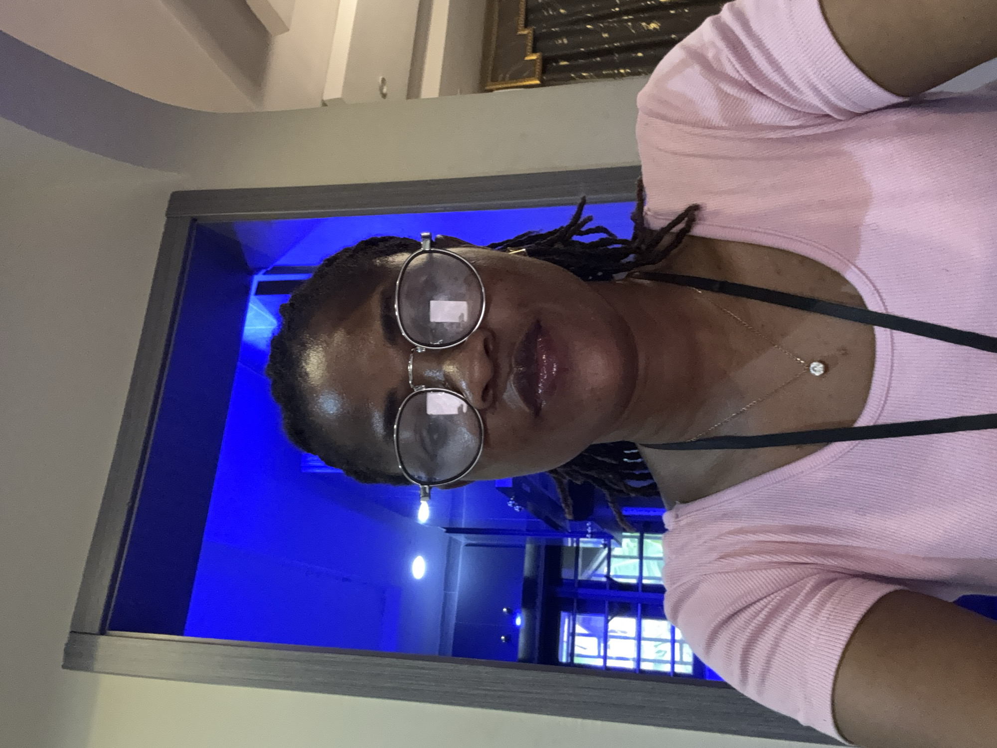 The media file shows a close-up, vertically oriented selfie of a person wearing a pink top and glasses. The subject has dark skin and is looking towards the camera, with their lips slightly parted. They are wearing round, silver-rimmed glasses and a delicate necklace with a small pendant. Their hair is styled in dark braids that fall over their shoulders. The background is dominated by a vibrant, electric blue light, suggesting an indoor setting with specialized lighting, possibly a studio or a themed room. The lighting creates a dramatic and somewhat surreal atmosphere. The image appears to be taken indoors, with what looks like a doorway or opening to the left, framing the subject against the blue background. The overall impression is one of a stylized portrait, emphasizing the subject's features against the striking backdrop. There is no visible text in the image, and the time of day and weather are not discernible.