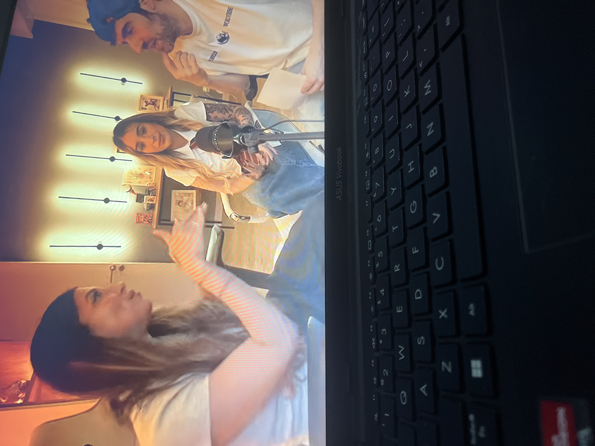 The image shows a laptop screen displaying a video call or broadcast featuring three people. On the left, a woman with long, blonde hair and a white t-shirt is speaking with her hands outstretched. In the center, a woman with blonde hair, wearing a white shirt with a tie and blue jeans, is seated and looking forward. To the right, a man wearing a blue cap and a white t-shirt is holding a piece of paper and appears to be speaking. The background of the broadcast shows a shelf with items and decorative lights. The laptop's keyboard is visible in the foreground. The location in the city of Santa Ana, Venezuela, is not indicated in the image.