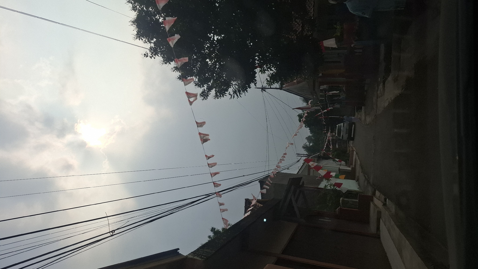 The image depicts a street scene in Sukasari, Indonesia, on an overcast day with the sun partially visible through the clouds. The street is lined with houses and trees. Red and white triangular flags are strung across the street, indicative of a national celebration. In the foreground, the dashboard of a vehicle is visible, suggesting the photo was taken from inside a car. Further down the street, a white car is parked, and a person in light blue clothing is walking away from it. Another person in light blue is standing near a building on the right side of the street. Power lines crisscross the sky. The overall impression is a residential area decorated for a festive occasion.