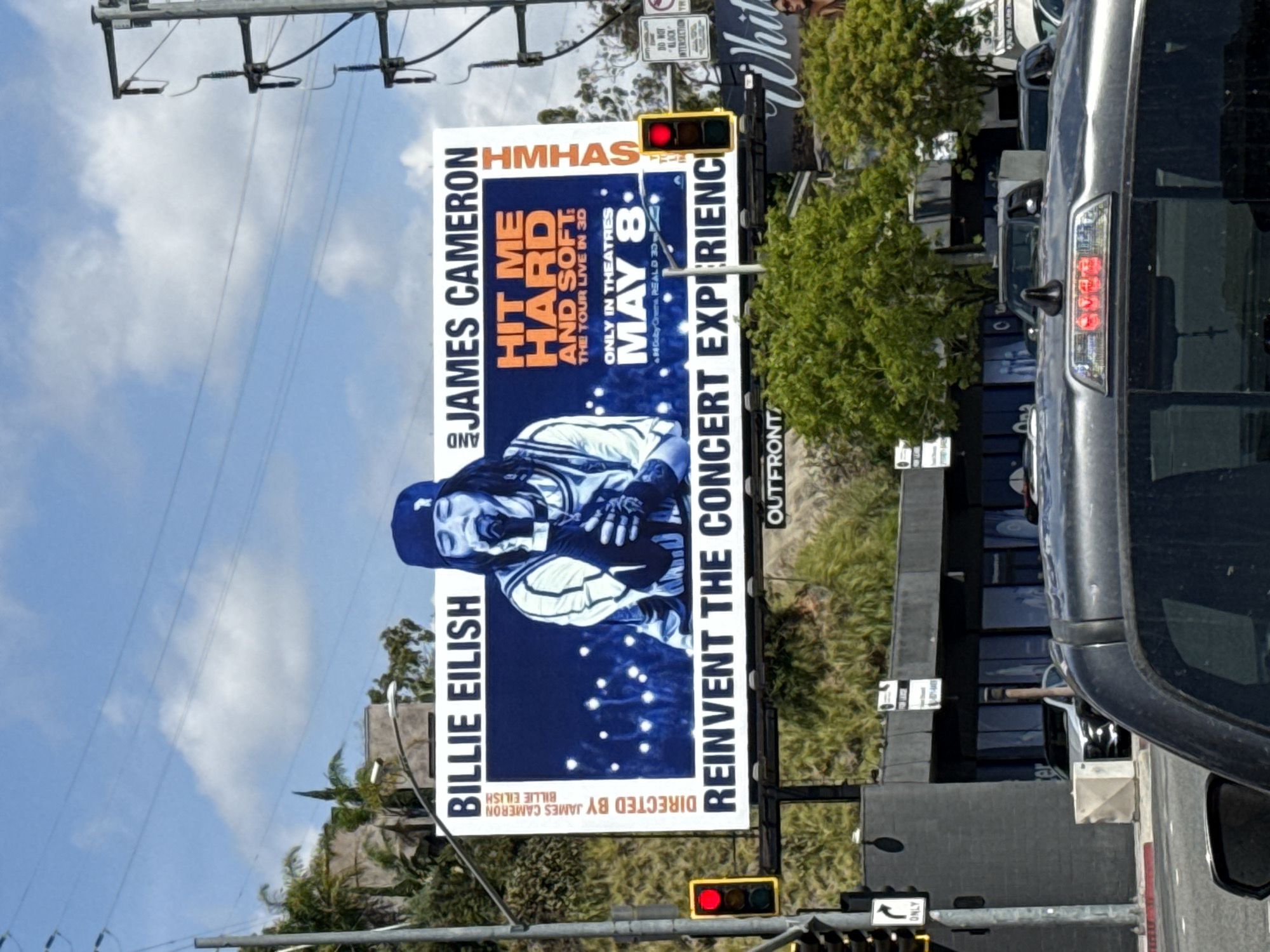 The image shows a billboard ad for Billie Eilish and James Cameron's "Hit Me Hard and Soft: The Tour Live in 3D", which will be in theatres May 8. The location is West Hollywood, United States of America, indicated by the "DO NOT BLOCK INTERSECTION" sign, and the billboard itself which promotes Outfront, a billboard advertising company.