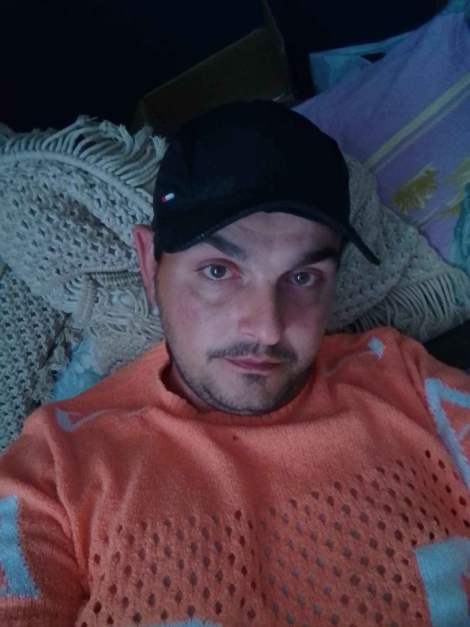 A man wearing a black baseball cap and an orange, open-knit sweater is lying down, looking at the camera. He has light brown eyes and a light mustache and stubble. The man is on a surface covered with a textured, cream-colored blanket with fringe. Behind him, other blankets are visible, one of which is purple with yellow stripes. The image appears to be taken indoors, likely in a residential setting.
