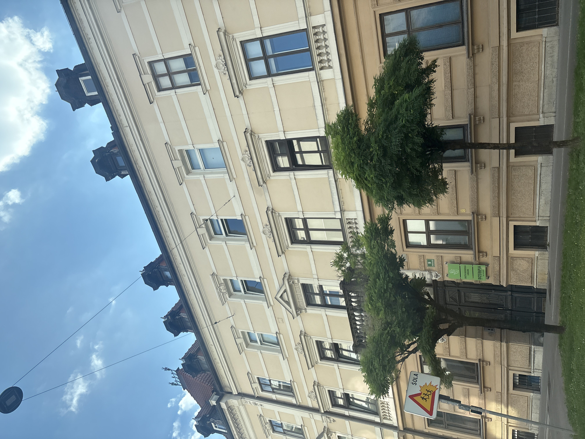 The image shows a street scene with an old, light yellow building featuring numerous windows and decorative elements. Two trees with full green foliage are positioned in front of the building, partially obscuring it. A traffic sign with a yellow triangle and images of children is visible on the right side of the frame. The sky is blue with some white clouds. The image is taken from a low angle, looking up towards the building and sky. The orientation of the image is rotated, with the building appearing on the left and the sky on the right. There are a few signs on the building, one of which appears to be green with white text, and another with the number "9". The text on the green sign is difficult to read but appears to include words like "DOSTOP" and "BIOTERAPIJA". The traffic sign contains the word "ŠOLA" in white text above red and yellow graphic representations of children. The lighting suggests it is daytime, likely sunny due to the clear sky and the way light falls on the building. The overall scene conveys a sense of an urban environment with architectural interest and greenery.