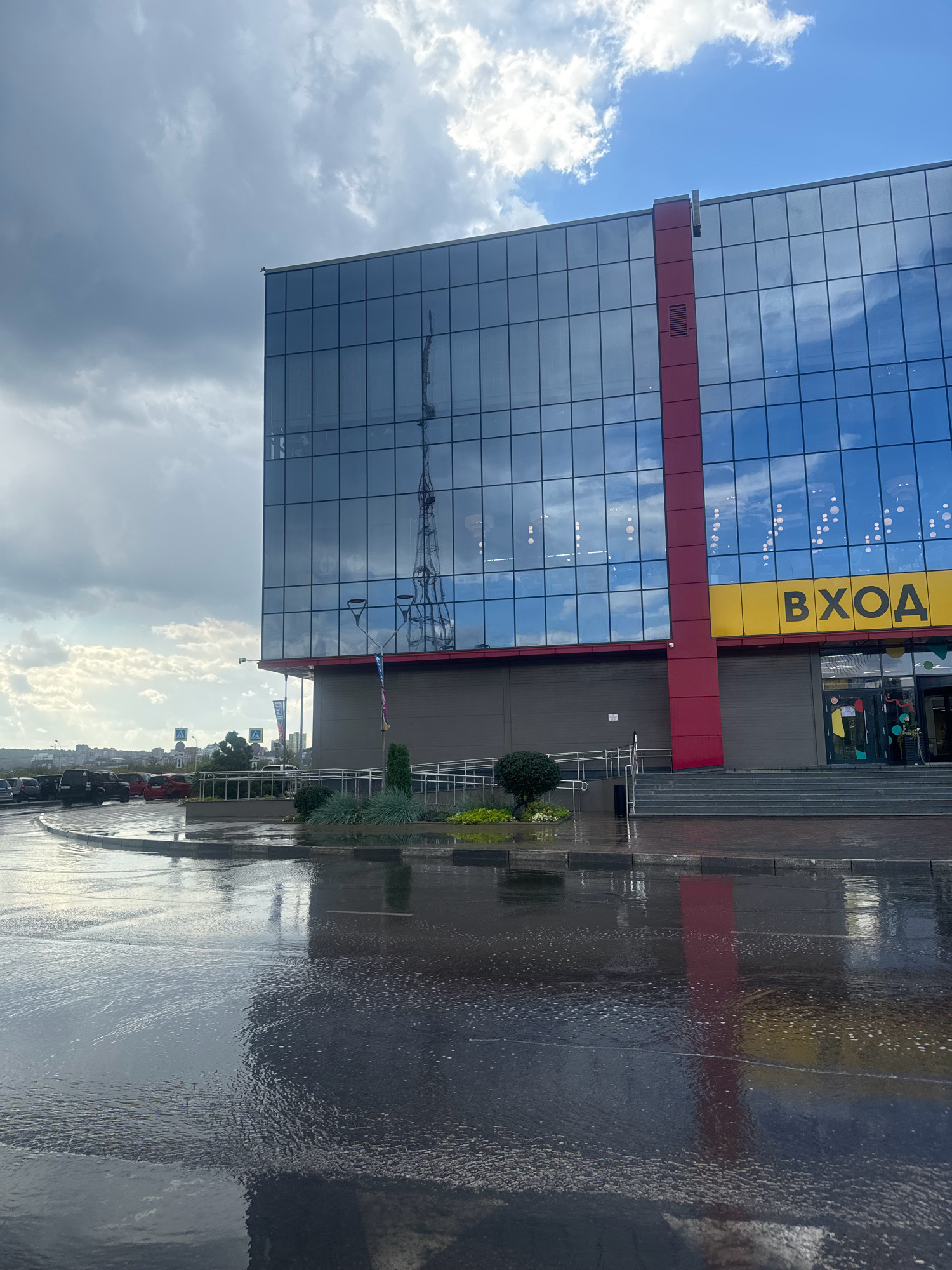 The image shows a modern, glass-fronted building with a red vertical accent. A large yellow sign with the Russian word "ВХОД" (meaning "ENTRANCE") is visible above the entrance. The building is surrounded by a wet paved area, suggesting it has recently rained or is currently raining. Several cars are parked in the background on the left. The sky is cloudy, with patches of blue sky visible. The reflection of a transmission tower is prominent in the glass facade of the building. Based on the architecture and the "ВХОД" signage, this is likely a commercial building or shopping center located in Irkutsk, Russia. The scene is captured from a low angle, looking towards the building.