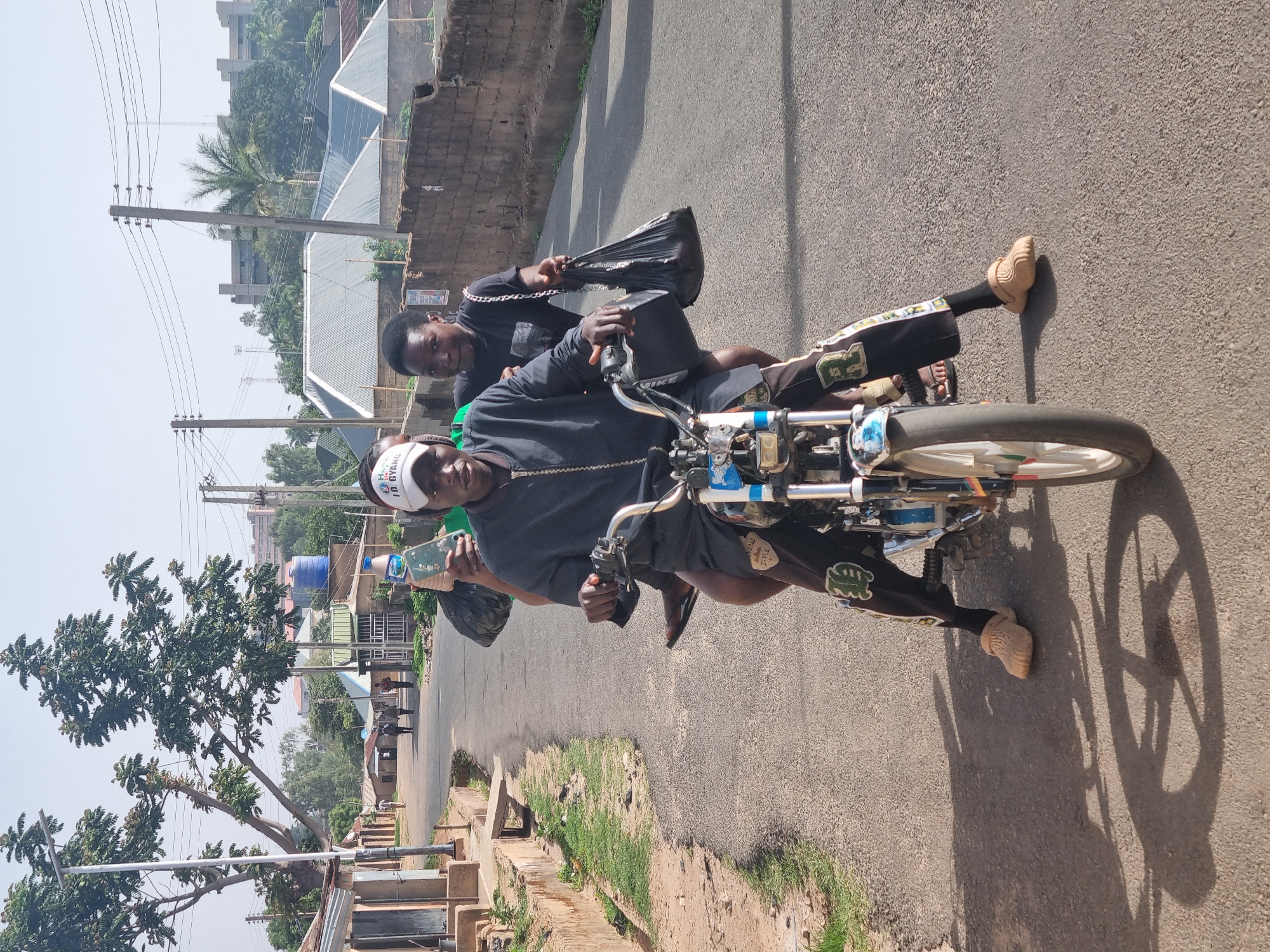 Two individuals are riding a motorcycle on a paved road. The rider wears a white baseball cap and dark clothing. The passenger, positioned behind, is also wearing dark clothing and carries a black plastic bag. The motorcycle is blue and white. The environment includes urban elements such as buildings, utility poles, and vegetation. The image is likely taken during daylight hours in Jos, Nigeria.