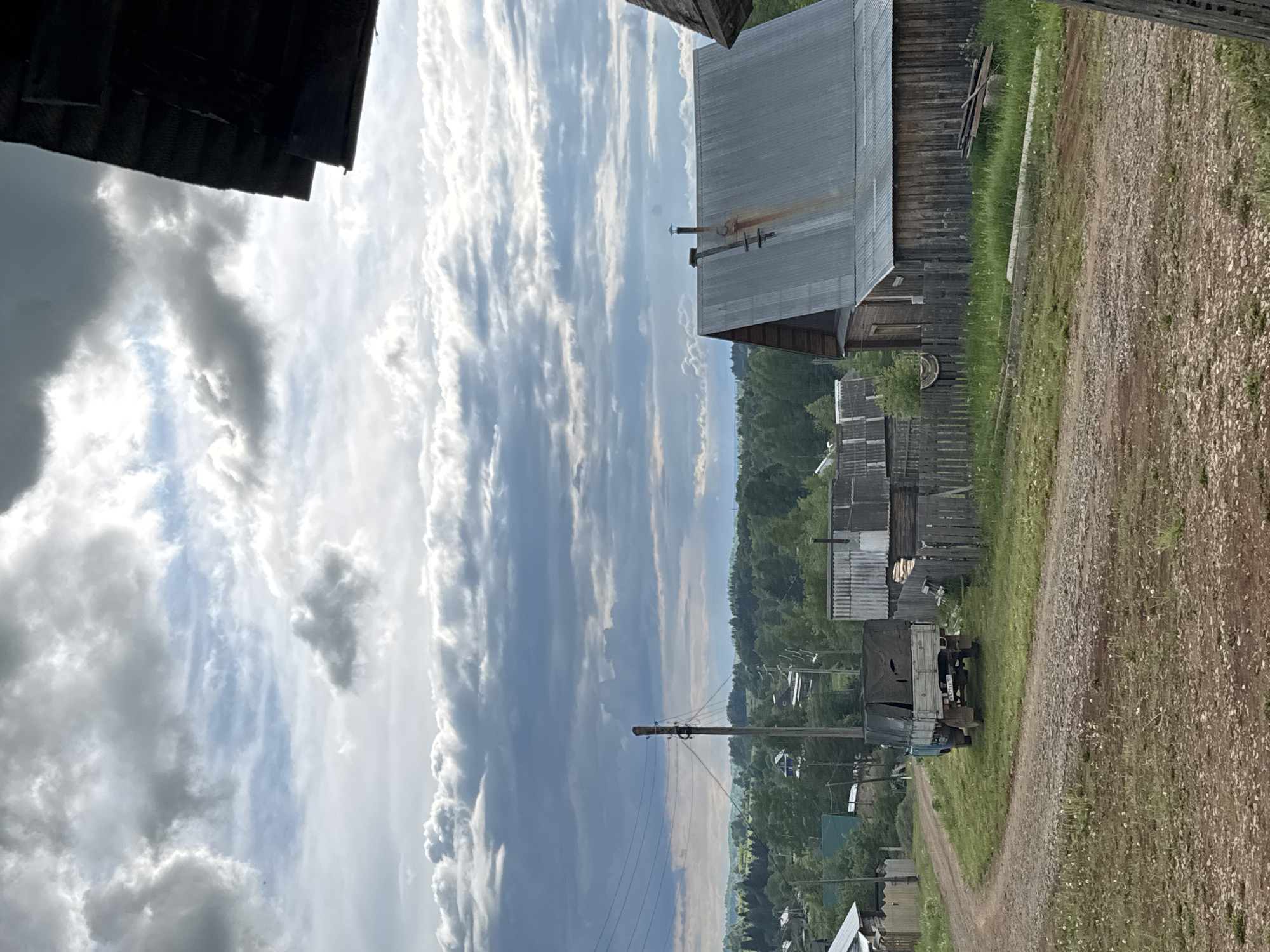 The image presents a panoramic view of a rustic village in Бородулино, Russia, under a dynamic, partly cloudy sky. A winding dirt road dominates the foreground and extends into the distance, flanked by traditional wooden houses.
On the right side of the road, several wooden buildings with corrugated metal roofs are visible, some showing signs of rust. Wooden fences separate the properties, and lush green grass grows abundantly alongside the path and around the structures. In the mid-ground, a utility pole with multiple power lines stands by the road. Further along, an older model truck, possibly a Soviet-era design, is parked; it has a light-colored cabin and a dark canvas cover over its bed. A license plate is visible on the rear of the truck, but the characters are indistinct.
Beyond the immediate village structures, the landscape slopes into a vast valley, densely covered with green trees and foliage, suggesting a forested or hilly terrain. Faint outlines of more distant buildings can be discerned in the valley. The sky is a prominent feature, filled with a mix of dramatic white and gray clouds, including long, streaky formations and larger, fluffy masses, with patches of blue sky peeking through. The scene is brightly lit, indicating daytime conditions. No people are visible in the image, contributing to a tranquil and serene rural atmosphere.