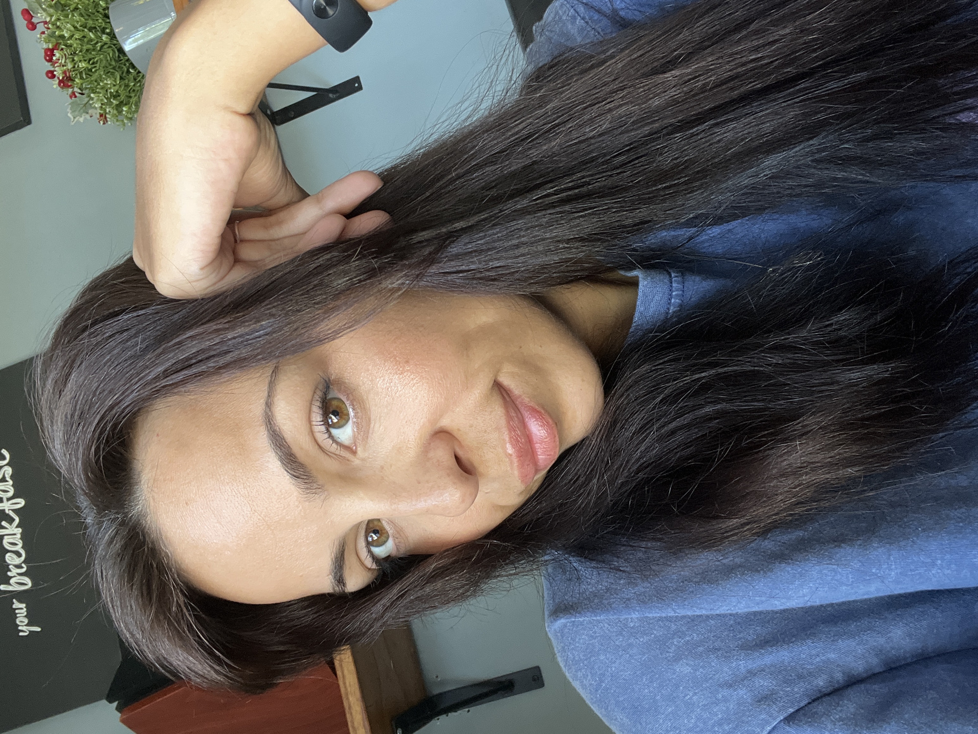A woman with long, dark hair and light brown eyes is looking at the camera. She is wearing a blue shirt and a black smartwatch. Her hand is near her ear. A small potted plant is visible in the background, along with a dark-colored wall and a wooden surface. The location in the city of Kuta Selatan, Indonesia is not discernible from the image.