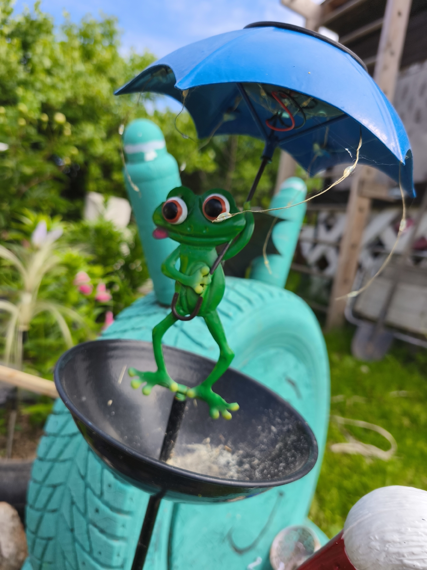 In Puchezh, Russia, a small, green, plastic frog figurine stands on a black dish. The frog holds a miniature blue umbrella, which is tilted open. The frog has large, bulging eyes with red rings and a pink tongue sticking out. It appears to be made of molded plastic and is mounted on a larger, teal-colored object that resembles a tire. The background is a blurred outdoor scene with green foliage and a bright blue sky. There are some indistinct objects in the background, including what appears to be a wooden structure. The overall impression is of a whimsical garden decoration.