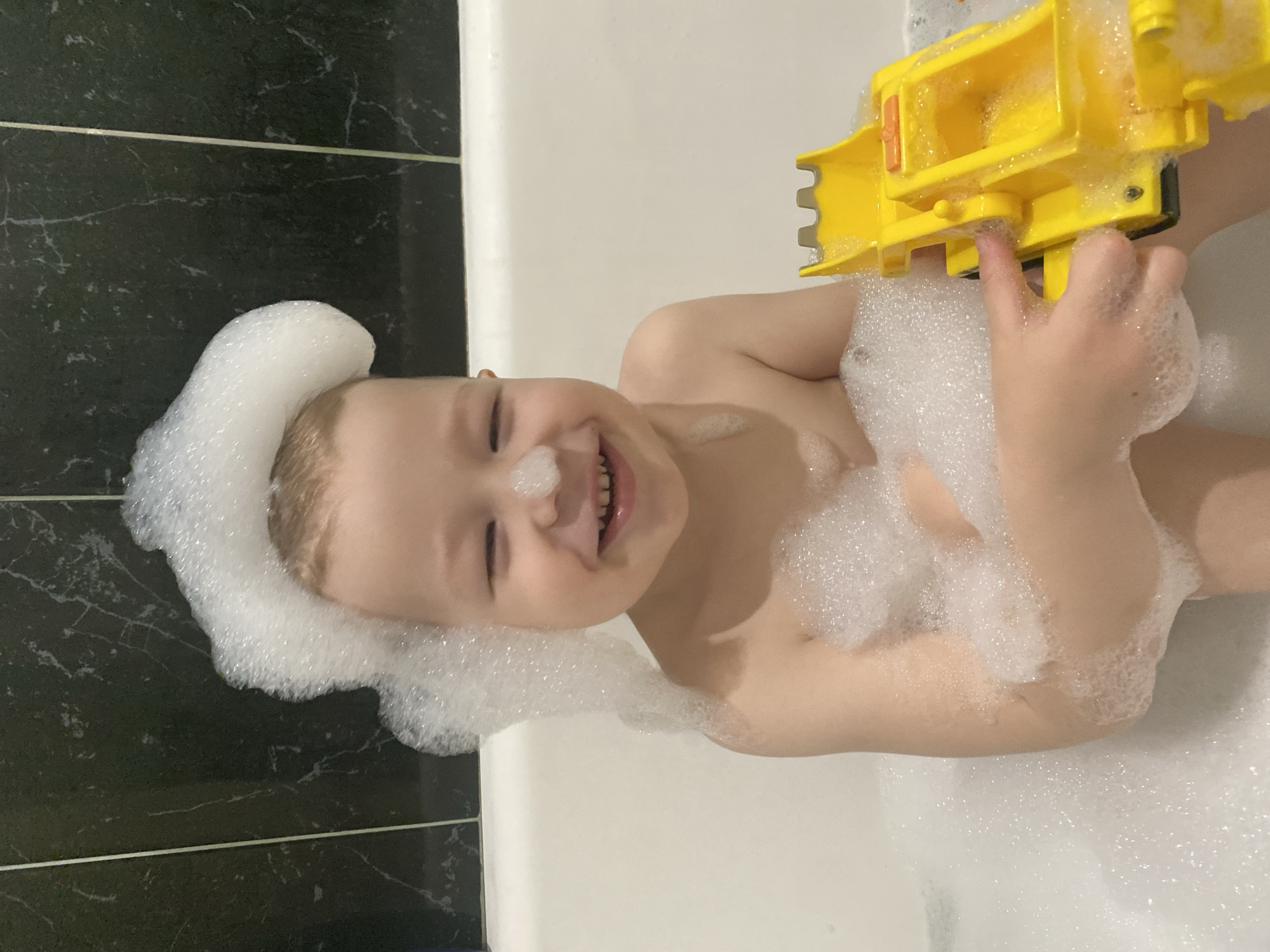 A child is in a bathtub filled with bubbles. The child has bubbles on their head and nose, and is holding a yellow toy truck. The bathroom has dark-colored tiles. This image does not contain enough information to determine whether or not it was taken in Sochi, Russia.