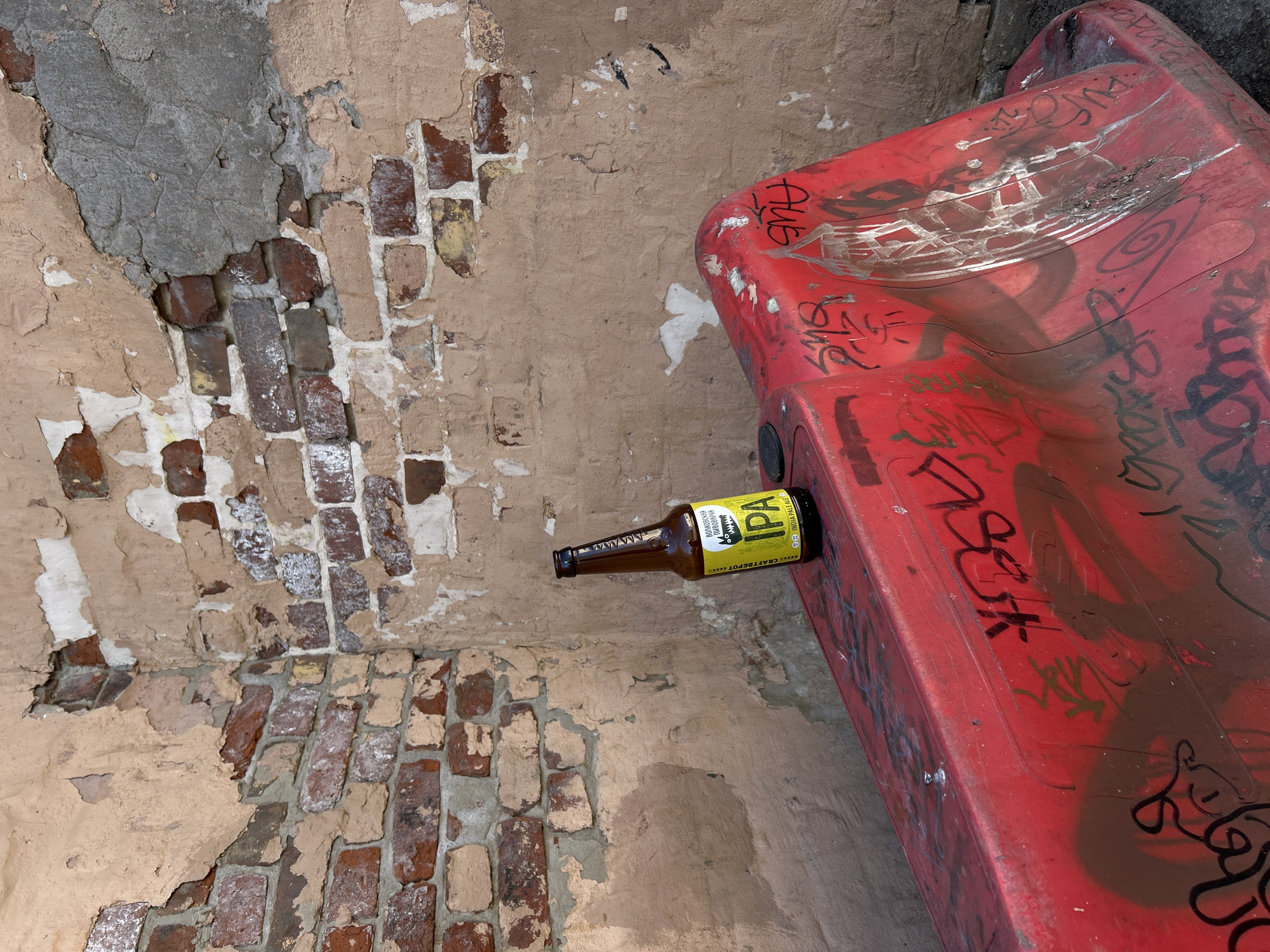 The image captures a ground-level or downward-looking perspective of an urban setting in Saint Petersburg, Russia, characterized by decay and neglect.
The scene is dominated by a distressed wall made of red and brown bricks, extensively covered by peeling and crumbling light brown plaster. Large sections of the plaster have fallen away, revealing the underlying brickwork and some patches of grey material, possibly older concrete or mortar. The texture of the wall is rough and uneven, showing signs of significant age and deterioration.
Leaning against this wall is a large, molded red object, likely made of plastic or fiberglass, which appears to have a curved, wavy surface, perhaps resembling a section of a slide or a stylized public barrier. This red object is heavily covered with various layers of graffiti, including tags and drawings in white, black, and brown spray paint. Visible graffiti includes stylized lettering and some discernible tags like "AUE".
Resting horizontally on the red object, near the corner where it meets the wall, is a brown glass beer bottle. Its yellow label is clearly visible. The label prominently displays "IPA" in large black letters, with additional text in Russian and English. The Russian text includes "ТРИУМФАЛЬНАЯ... ВОЛКОВСКАЯ ПИВОВАРНЯ" (Triumphal... Volkovskaya Brewery), and other text such as "CRAFTDEPOT" and "INDIA PALE ALE".
No human activity or individuals are present in the image. The lighting suggests it is daytime, with no direct sunlight indicating a specific time. The overall impression is one of urban grittiness, abandonment, and the passage of time on man-made structures.