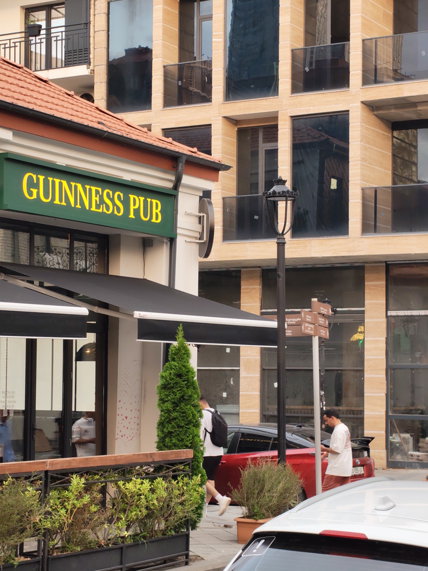 The image captures a street scene in Batumi, Georgia, featuring the "GUINNESS PUB" with a prominent green sign and yellow lettering. The pub has a red-tiled roof and outdoor seating areas with black awnings. Adjacent to the pub is a modern building with beige stone cladding and large reflective windows, characteristic of contemporary architecture.
In the foreground, a small evergreen tree stands to the right of the pub entrance, and a planter box with green shrubs is visible along the sidewalk. A vintage-style black lamppost with ornate detailing stands on the sidewalk. Several signs on a pole near the lamppost appear to be directional.
Two men are present in the scene. One man, wearing a white t-shirt, black shorts, and carrying a black backpack, walks away from the camera towards the right. Another man, also in a white t-shirt and light brown shorts, is standing near the lamppost, looking down as if interacting with a phone or object in his hand.
Two cars are parked on the street: a red car with a spoiler and a silver car partially visible in the lower right corner. The environment suggests an urban setting with a mix of traditional and modern architectural styles.