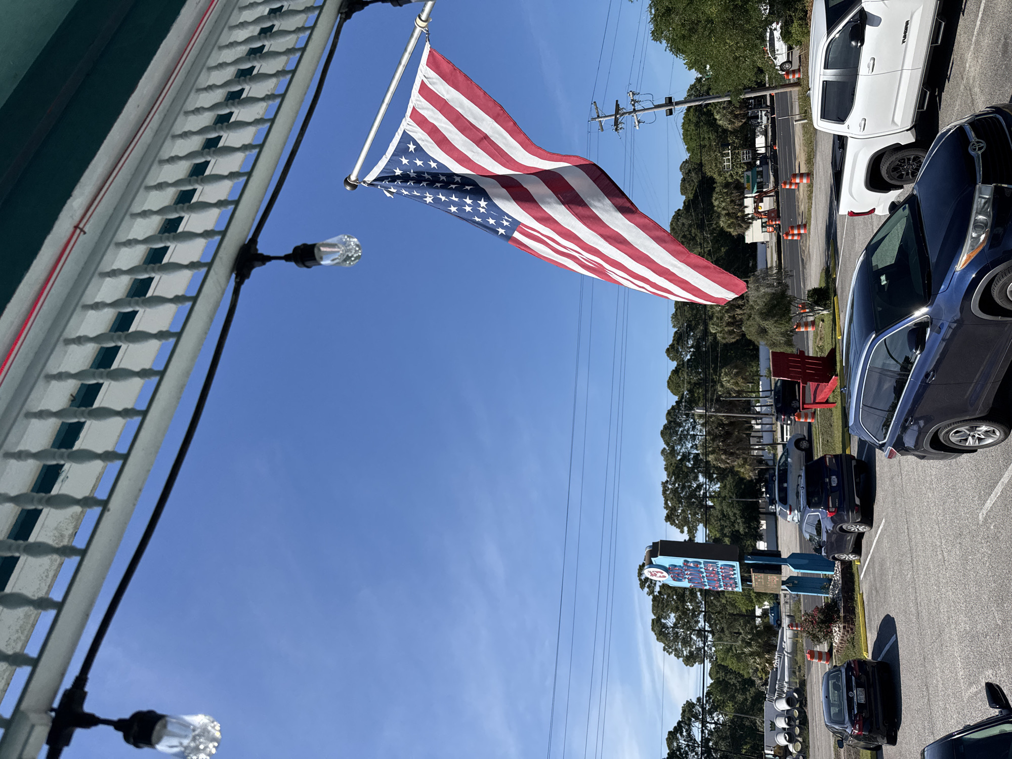 A slightly rotated, eye-level shot captures a vibrant scene on a clear, sunny day in Garden City Beach, South Carolina. An American flag, rippling in the wind, is prominently displayed, affixed to a building featuring white decorative latticework and vintage-style globe lights. A utility pole with numerous power lines is visible against the clear blue sky.
The foreground reveals a parking lot filled with a variety of vehicles, including a dark blue SUV and a white pickup truck, among others. Beyond the parking area, a glimpse of street is visible, marked by orange traffic cones, suggesting ongoing construction or road maintenance.
Further in the background, a large, colorful sign stands amidst lush green trees and foliage, providing a distinct landmark in this coastal town. The composition is a blend of patriotism, everyday life, and civic activity in a typical American setting.