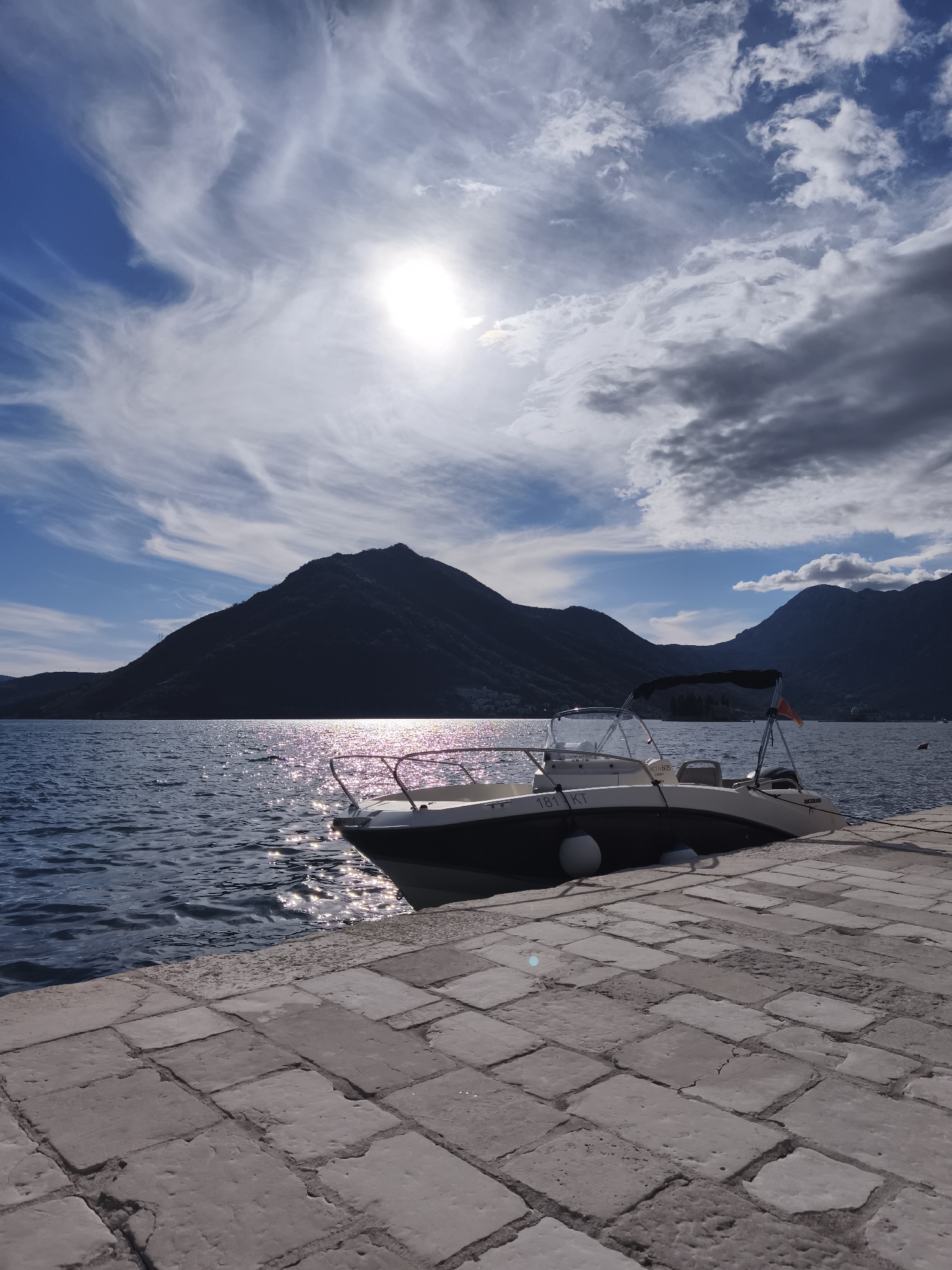 The image shows a sunny day at a waterfront location, likely in the city of Unknown, Unknown, with a boat docked alongside a stone pier. The sky is partially cloudy, with the sun shining brightly through the clouds. The calm water reflects the sunlight, creating a sparkling effect.
The boat is a small motorboat, with a white hull and a partially visible license plate with the inscription "181 KT". It is tied to the pier.
In the background, there are dark mountains, suggesting a coastal setting. The pier is constructed of large stone blocks.