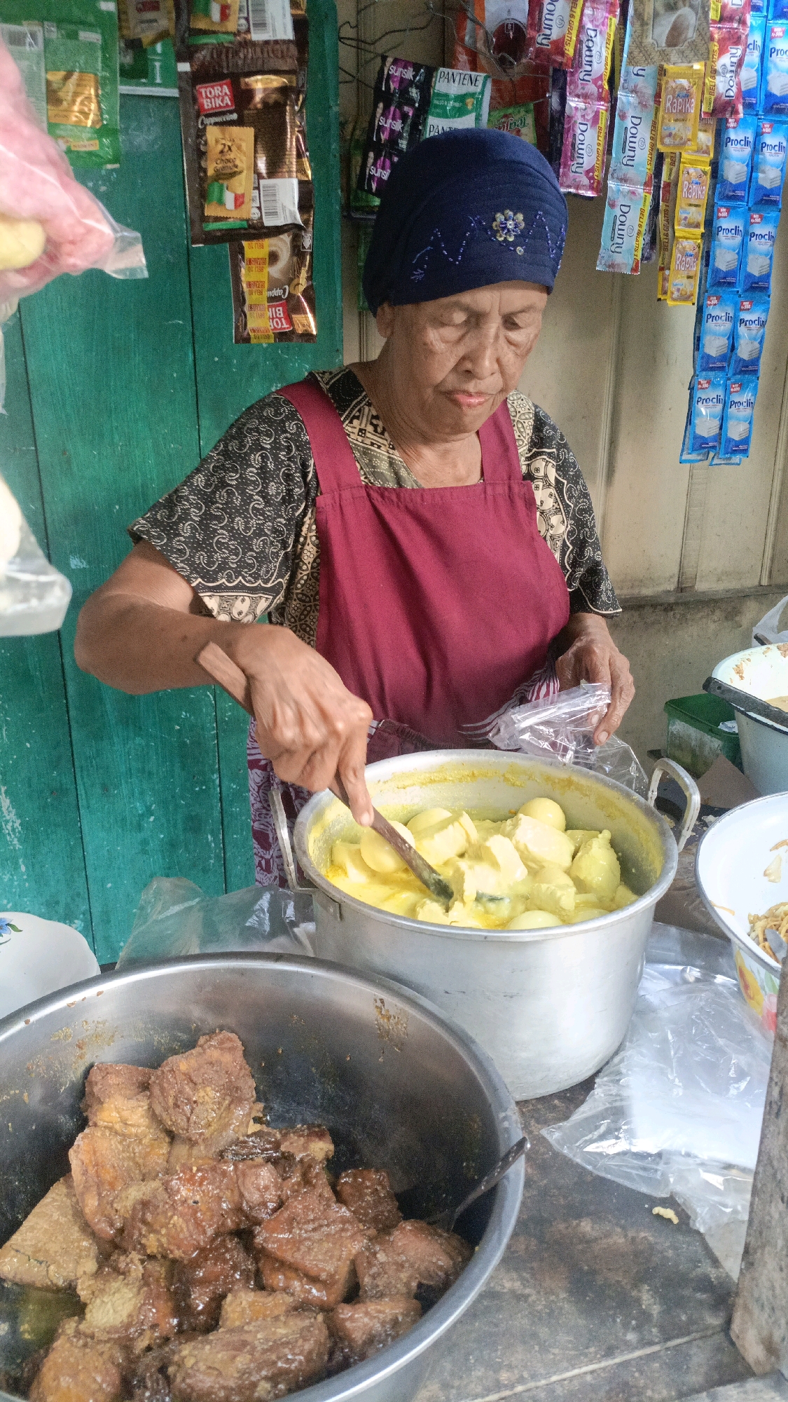 An elderly woman wearing a navy blue headscarf and a maroon apron is standing behind a market stall in Salatiga, Indonesia, preparing food. She is holding a wooden spoon and stirring a pot filled with a yellow curry-like dish containing what appear to be hard-boiled eggs and pieces of tofu or jackfruit. To her left, a large metal bowl contains chunks of dark brown, saucy meat, possibly beef or chicken. The stall has a turquoise wooden wall behind it and is adorned with various packaged goods hanging from racks, including snacks and cleaning supplies. The overall setting suggests a street food vendor in a local market.
