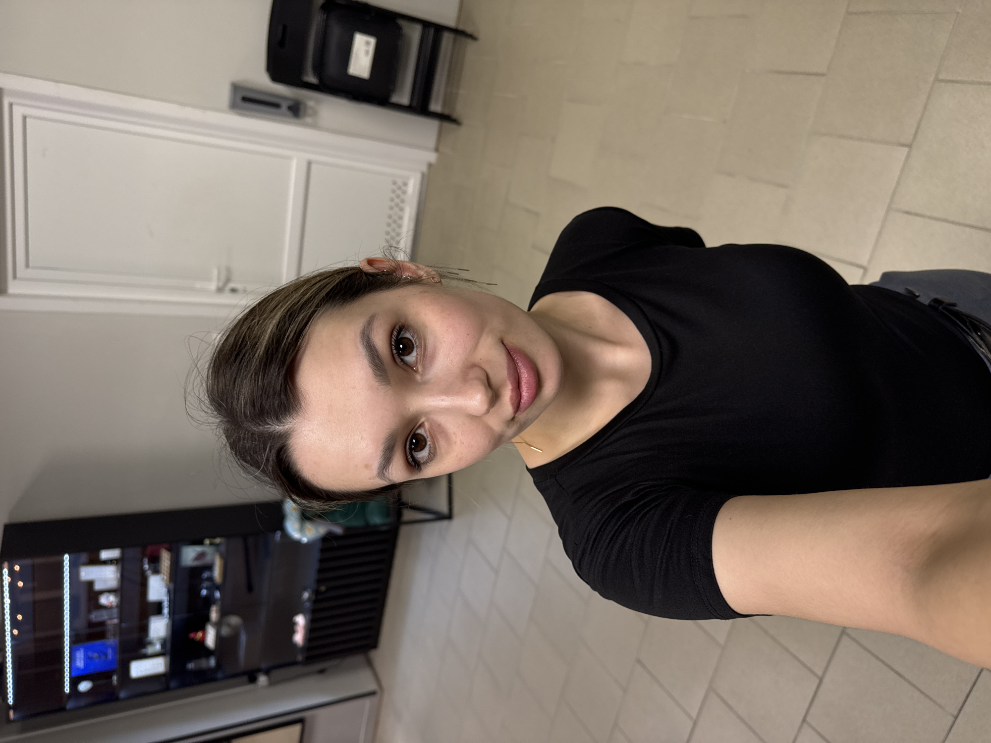 An upright selfie captures a young woman standing indoors, looking directly at the camera with a slight, gentle smile. Her dark brown hair, with subtle lighter highlights, is pulled back, revealing her face. She has large brown eyes and naturally pink, full lips. She wears a plain black long-sleeved or three-quarter-sleeved top and dark bottoms, possibly jeans or trousers, with a dark belt. A delicate gold bar necklace rests on her collarbone, and small earrings are visible in her right ear, along with a few hairpins keeping her hair in place.
The background suggests an interior commercial or public space. The floor is covered with light-colored, square tiles. Behind her, a plain white door with simple molding details is visible. To her right, a tall, dark display case or shelving unit showcases various blurred items, possibly products, and features a vertical strip of bright LED lights on its left edge. Higher up in the background, near the ceiling, are a black rectangular object (possibly an overhead projector, speaker, or part of an HVAC system) and a horizontal vent. The overall lighting is artificial and bright, typical of an indoor setting, with some shine on her forehead and nose indicating a light source. No visible text is discernible in the image.