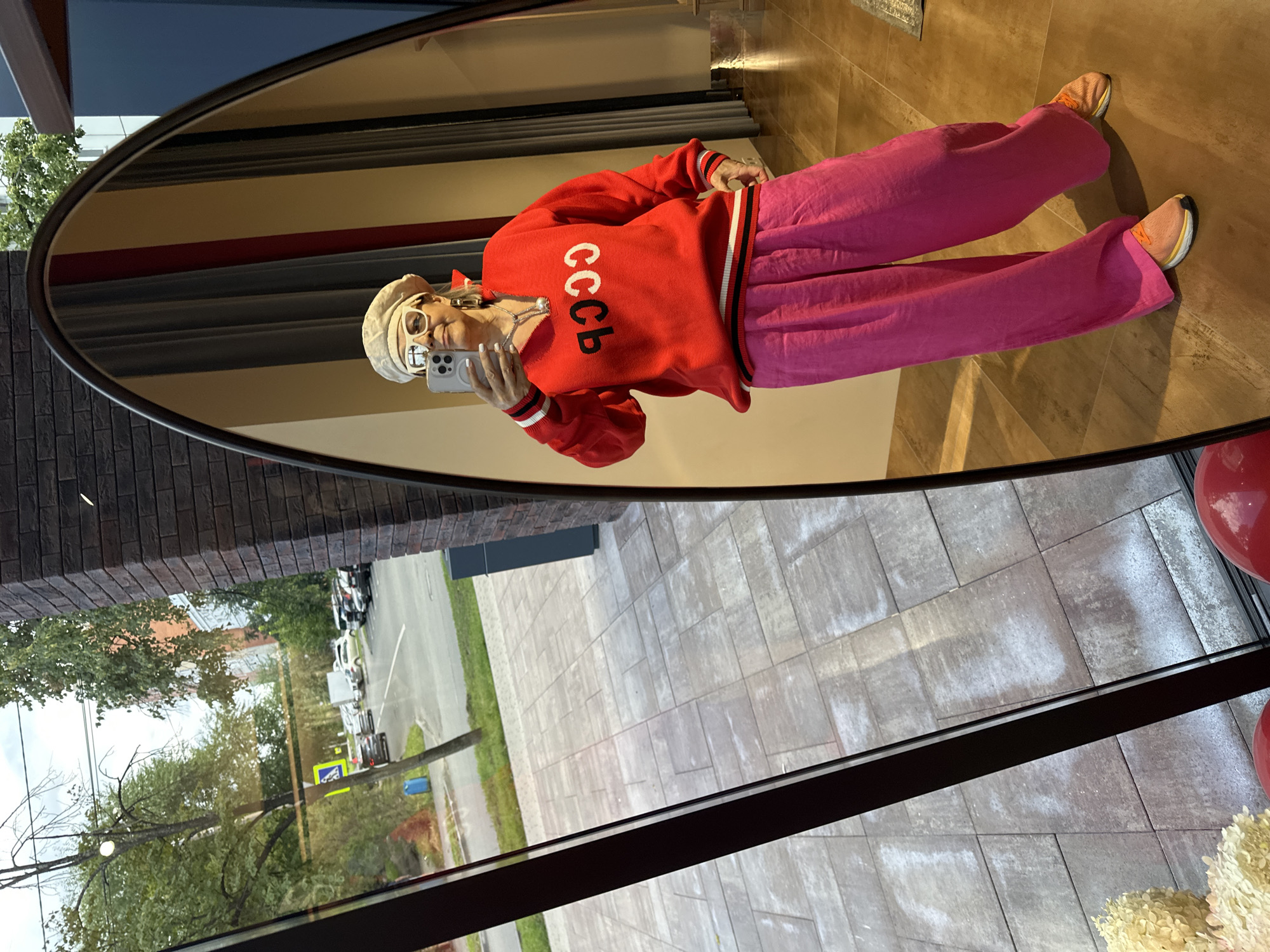 A woman, seen in a reflection in a large oval mirror, stands indoors, taking a selfie with her phone. She is wearing a beige beret, large white-rimmed glasses, a red V-neck sweater with "CCCP" in black and white lettering, and bright pink wide-leg pants. She is also wearing a silver necklace and hoop earrings. Her feet are clad in orange athletic shoes. The background visible in the mirror includes striped drapes and a reflection of the interior of what appears to be a store. The mirror is positioned to reflect an outdoor scene visible through a window or glass facade to the left, showing a street with parked cars, trees, and buildings. The floor in the foreground of the reflection is tiled.