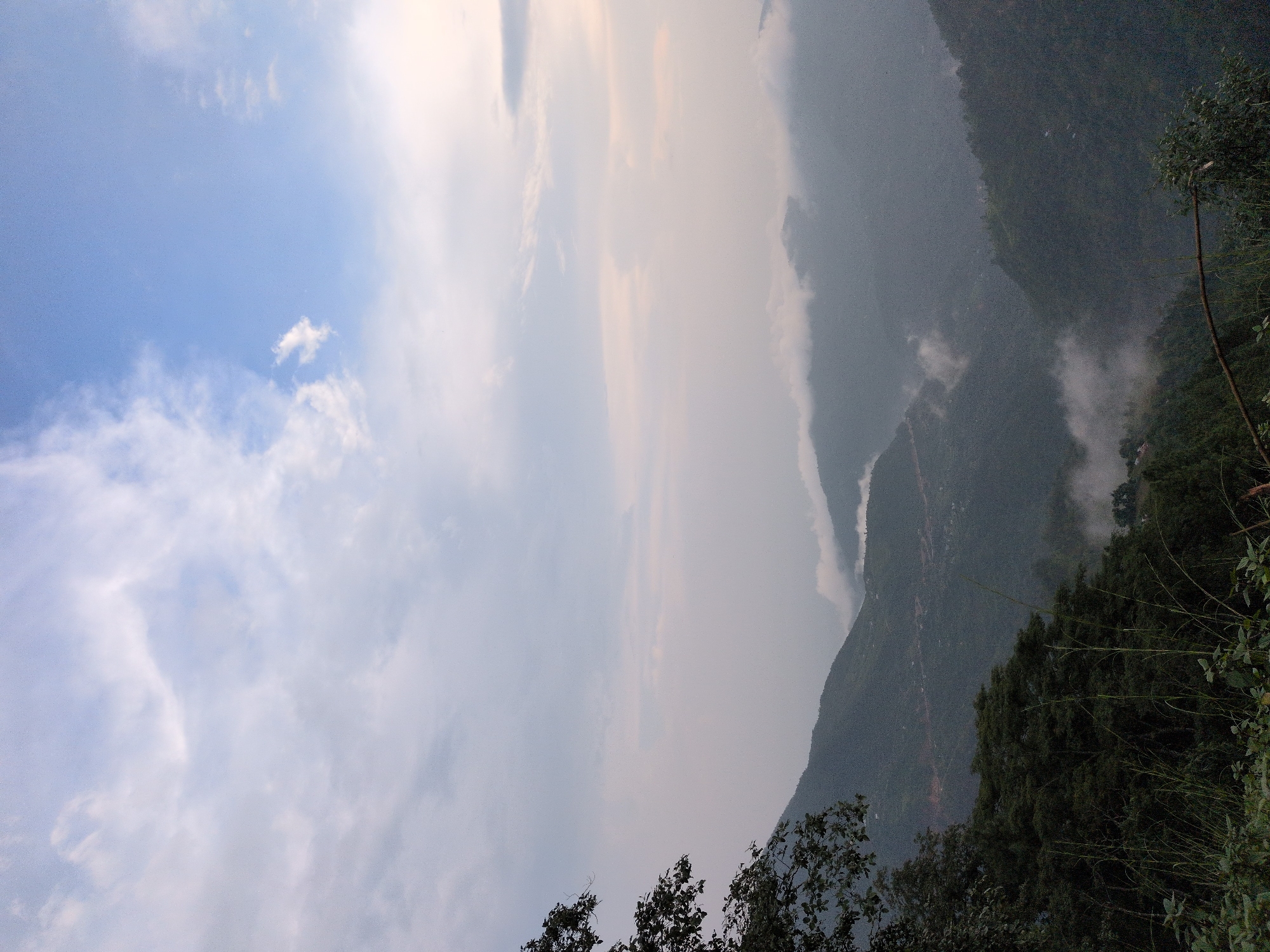 The image depicts a serene mountainous landscape near Kapurkot, Nepal. The sky is a gradient of pale blue, transitioning to a soft white near the horizon, with wispy clouds scattered across its expanse. In the foreground, lush green foliage and tall grasses frame the lower portion of the scene. Dominating the middle ground and background are rolling hills and mountains, layered in varying shades of deep green and muted grey due to atmospheric haze. A winding road is visible etched into one of the hillsides, suggesting human presence. Patches of mist and low-lying clouds drift through the valleys, adding a sense of depth and ethereal beauty to the mountainous terrain. The overall impression is one of natural tranquility and the vastness of the mountain environment.
