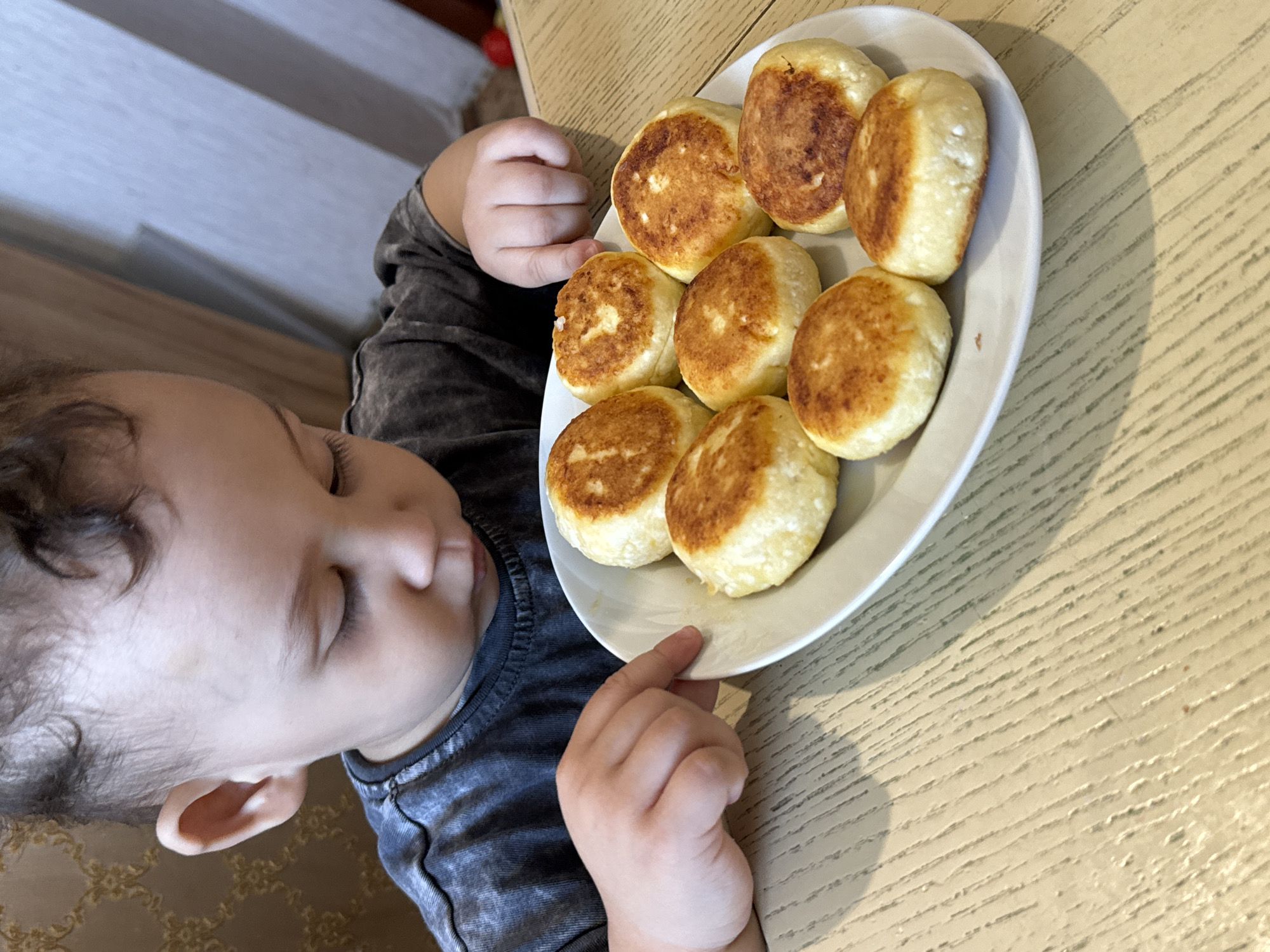 A young child lies beside a plate of golden-brown syrniki on a beige table with a wood grain pattern. The child, wearing a gray shirt, appears to be sleeping or resting their head near the plate. The plate contains nine syrniki, which are evenly spaced and appear freshly cooked. The photograph was taken indoors, presumably in a home in Chaikovsky, Russia, as specified. There is nothing present to further confirm its location or other surrounding environmental conditions.