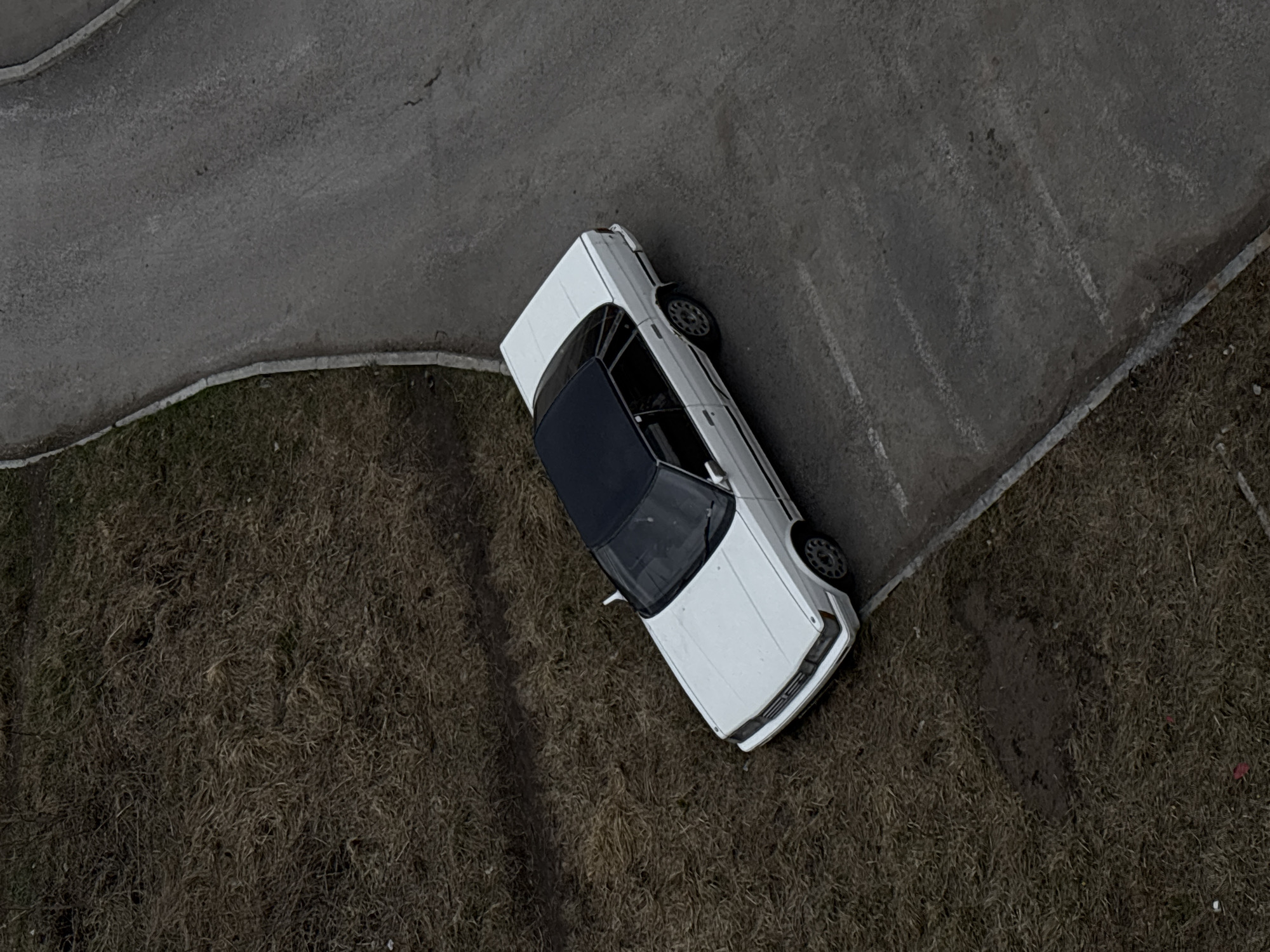 An aerial view depicts a white sedan with a black roof parked partially on a paved road and partially on a patch of dormant grass. The road surface appears gray and possibly cracked. The surrounding area is a mix of pavement and dry vegetation. The location is Perm, Russia.
