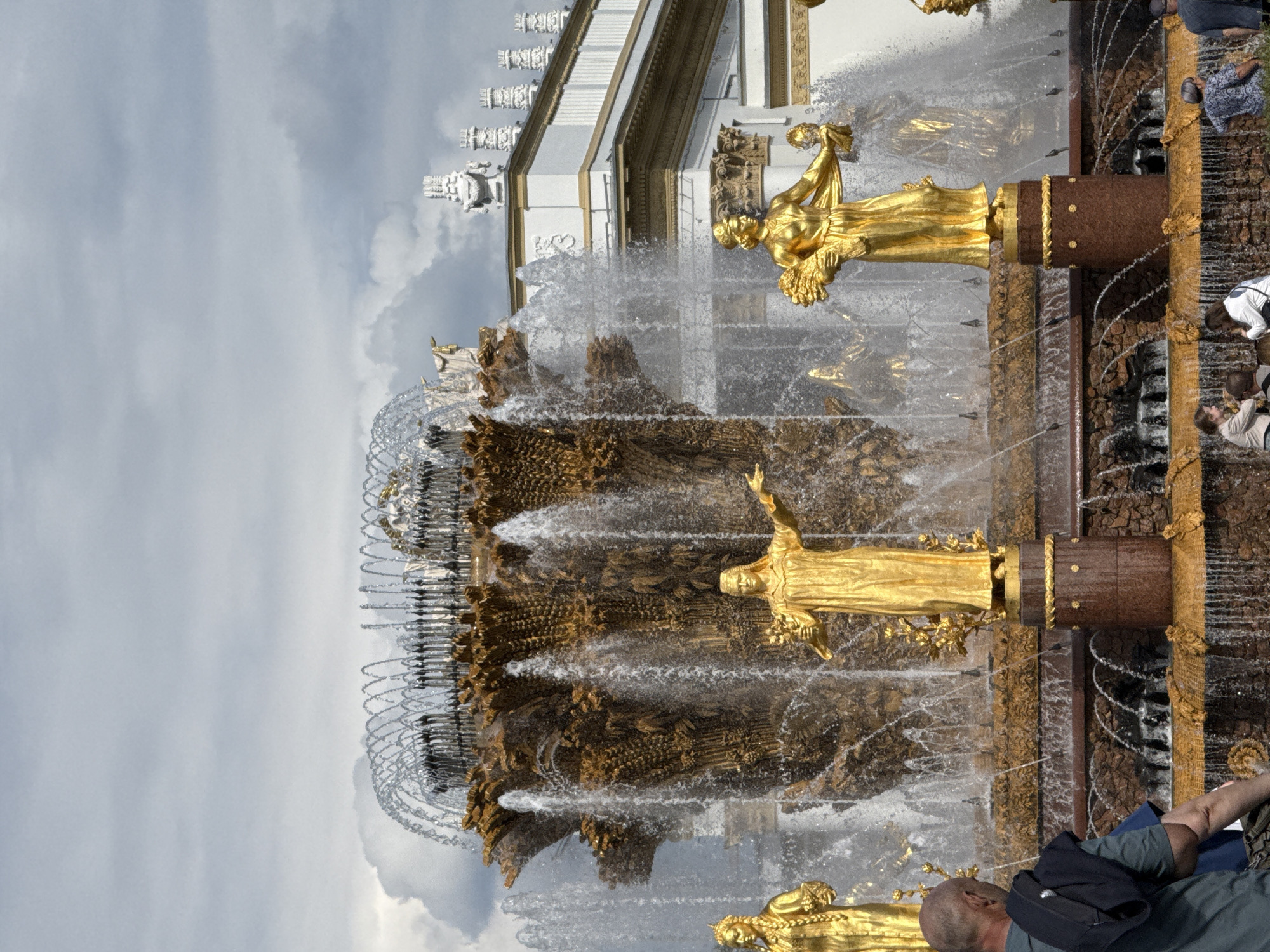 This media file captures a majestic fountain scene in Moscow, Russia. The central subjects are a series of ornate golden statues, depicting female figures, integrated into a multi-tiered fountain. Water cascades down the golden structures and flows into pools below, creating a dynamic and lively atmosphere. The background reveals part of a grand building with classical architectural elements, including columns and decorative friezes, suggesting a significant public space or historical landmark.
Several people are visible in the foreground and along the edges of the fountain. They appear to be tourists or visitors, observing the fountain and enjoying the ambiance. Some are leaning over to get a closer look or take pictures, while others are seated or standing, their attention drawn to the water display. One person in the lower right corner is seen wearing a backpack and looking towards the fountain.
The weather appears overcast, with a cloudy sky visible in the upper portion of the frame. This suggests a moderate temperature, typical for a day when outdoor fountains are operational. The time of day is not explicitly indicated but appears to be daytime due to the lighting. The overall mood conveyed is one of awe and appreciation for the artistry and grandeur of the fountain and its surroundings. The golden statues, combined with the flowing water and impressive architecture, create a visually striking and memorable scene. No visible text is present in the image.