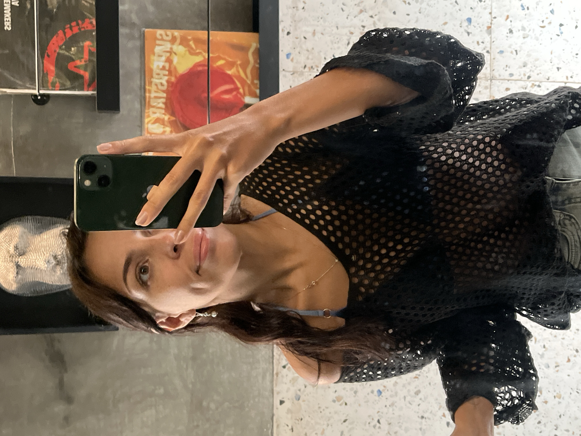 A woman takes a mirror selfie in an indoor setting, visible from the chest up. She holds a dark green/black iPhone with three camera lenses in her right hand, which partially obscures her face. She has dark brown, wavy hair and looks directly into the camera/mirror with a neutral expression and slightly parted lips. She wears a black mesh top with a distinct hole pattern, under which a light blue or grey bra strap is visible. Accessories include a delicate silver necklace and a dangling earring with a pearl.
The background, reflected in the mirror, shows an interior space with a terrazzo-style floor. On a shelf behind her, two items resembling album covers or books are reflected. The one on the left is dark with a red graphic and reversed text that appears to read "SREKAMEW" (likely "WEMAKERS" or "REMAKERS"). The item on the right is orange with a prominent red circular graphic, possibly a drum or speaker, and some indistinct reversed text. To the left of the woman's head, a light-colored, intricately patterned object, possibly a piece of coral or a natural sculpture, is reflected. The setting suggests a casual, possibly artistic or home environment.