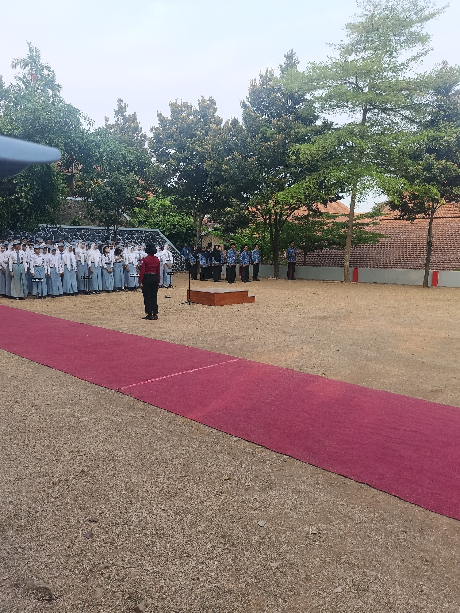 In Pengkol, Indonesia, a group of students, likely in uniform, stand in formation on a sandy ground. They are arranged in neat rows, facing a red carpet that stretches across the foreground. A single individual, possibly an instructor or leader, stands on the red carpet, facing the students. Behind the students, a stone wall is visible, and beyond that, trees and buildings with tiled roofs. The sky is overcast, suggesting a neutral lighting condition. The scene appears to be an outdoor event or ceremony taking place on the grounds of a school or institution.