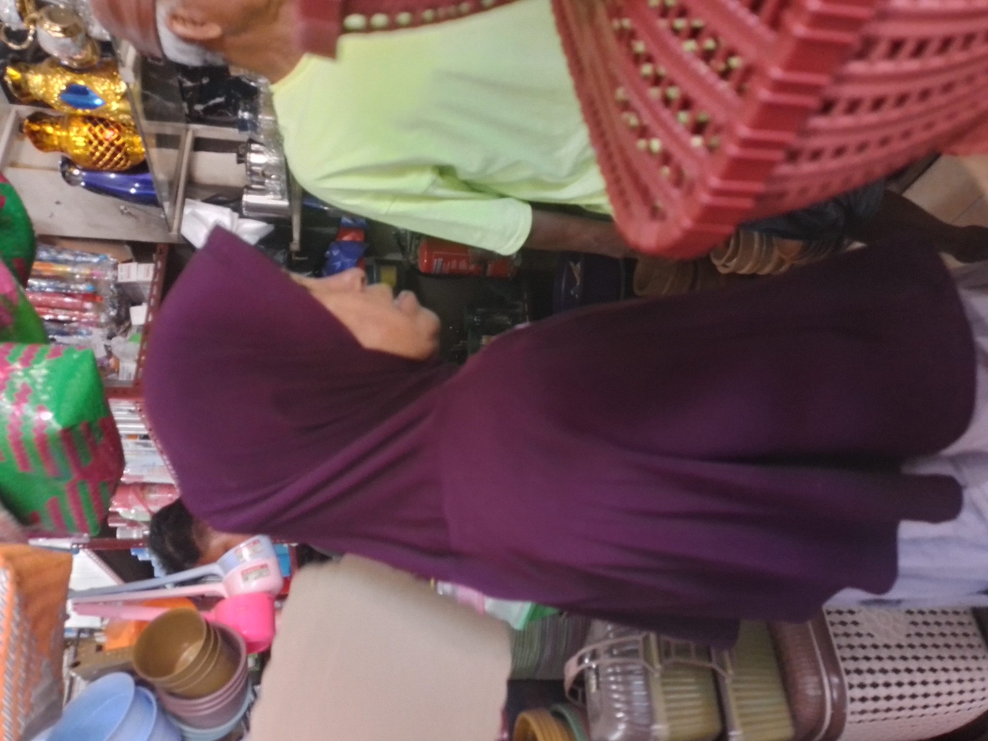 The image shows a market stall in an indoor setting, possibly a market or shop. In the foreground, a person wearing a purple hijab is partially visible, with their face turned to the left and their mouth slightly open. To the right of the person in the hijab, another person is visible wearing a light green t-shirt and a brown hat, with their head tilted down. They appear to be looking at something below the frame.
The background of the image is filled with various objects. On shelves, there are decorative items, including golden pineapple-shaped figures, blue vases, and a white teapot. Below the shelves, there are metal pitchers and cups. In the left and center of the image, various household items are displayed, including plastic bowls of different colors, brushes with pink and blue handles, and woven baskets. A red plastic basket with a patterned interior is prominently placed in the lower right portion of the image. The overall environment suggests a busy marketplace where goods are being sold.