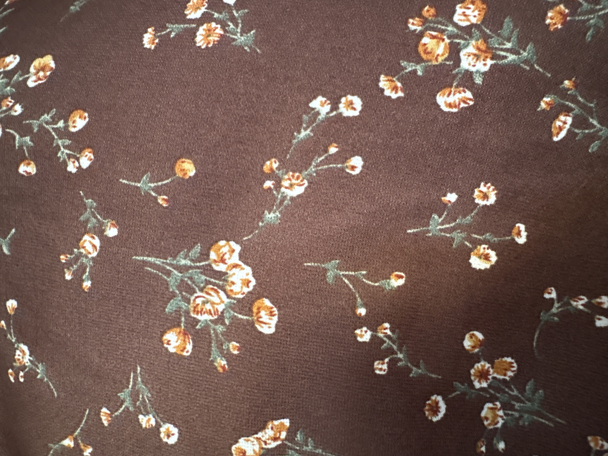 A close-up, overhead shot shows a brown fabric with a floral pattern. The flowers are small, with white and orange petals, and green stems and leaves. The pattern is scattered across the fabric, with no specific arrangement. The lighting is even, and there are no shadows, suggesting the fabric is not in direct sunlight. The texture of the fabric appears to be soft, possibly cotton or a similar material. There are no people or animals visible in the image.