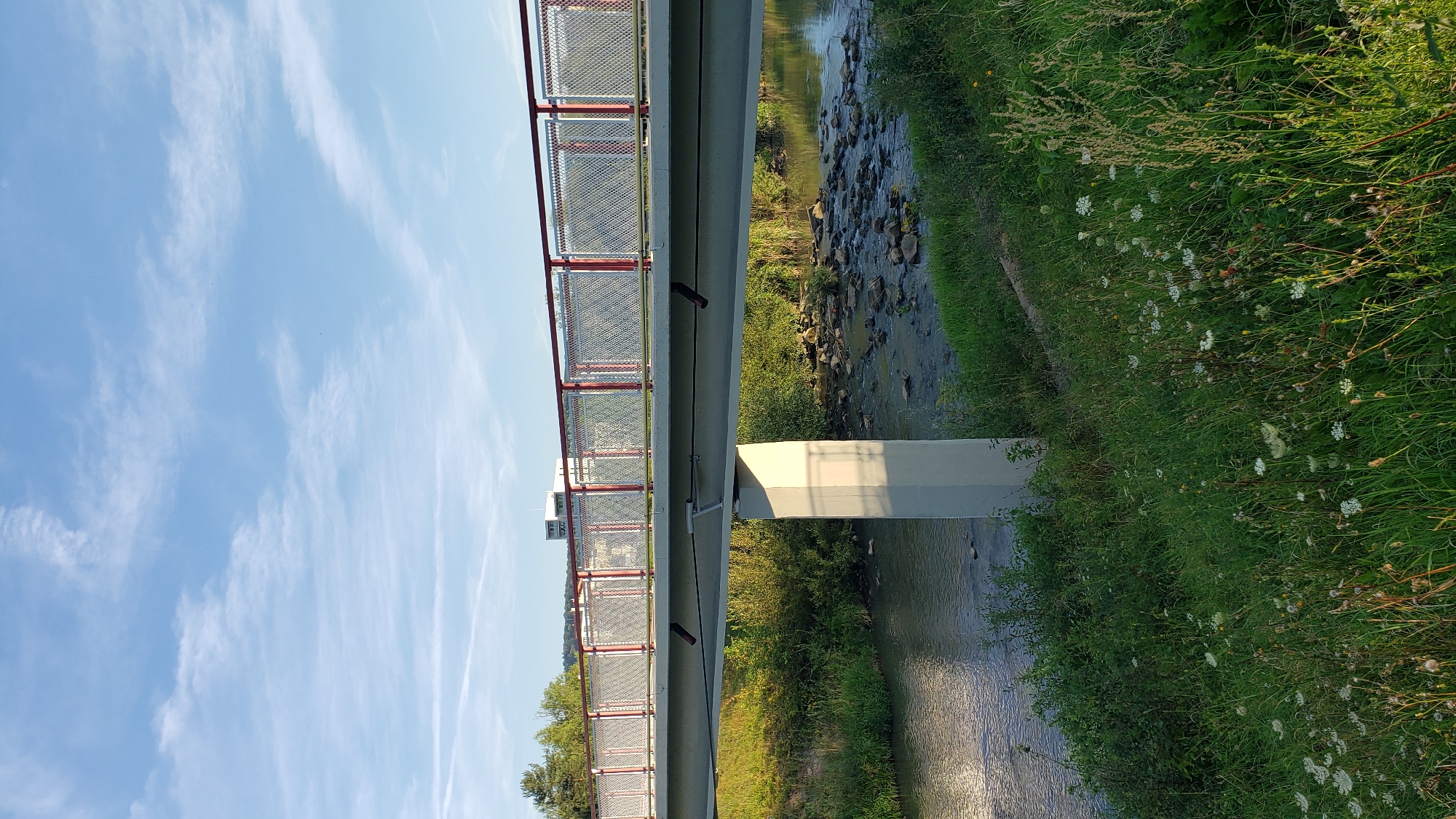 The image shows a pedestrian bridge over a small river, likely in the city of Prešov, Slovakia. The bridge has a concrete deck and is supported by a single concrete pillar in the middle of the river. The railings are made of metal with a red top and white mesh infill. The riverbed is rocky, and the water is relatively shallow. Lush green grass and wildflowers grow along the riverbanks. In the background, trees and a few buildings are visible. The sky is blue with scattered white clouds, suggesting it is daytime. The scene appears peaceful and natural.