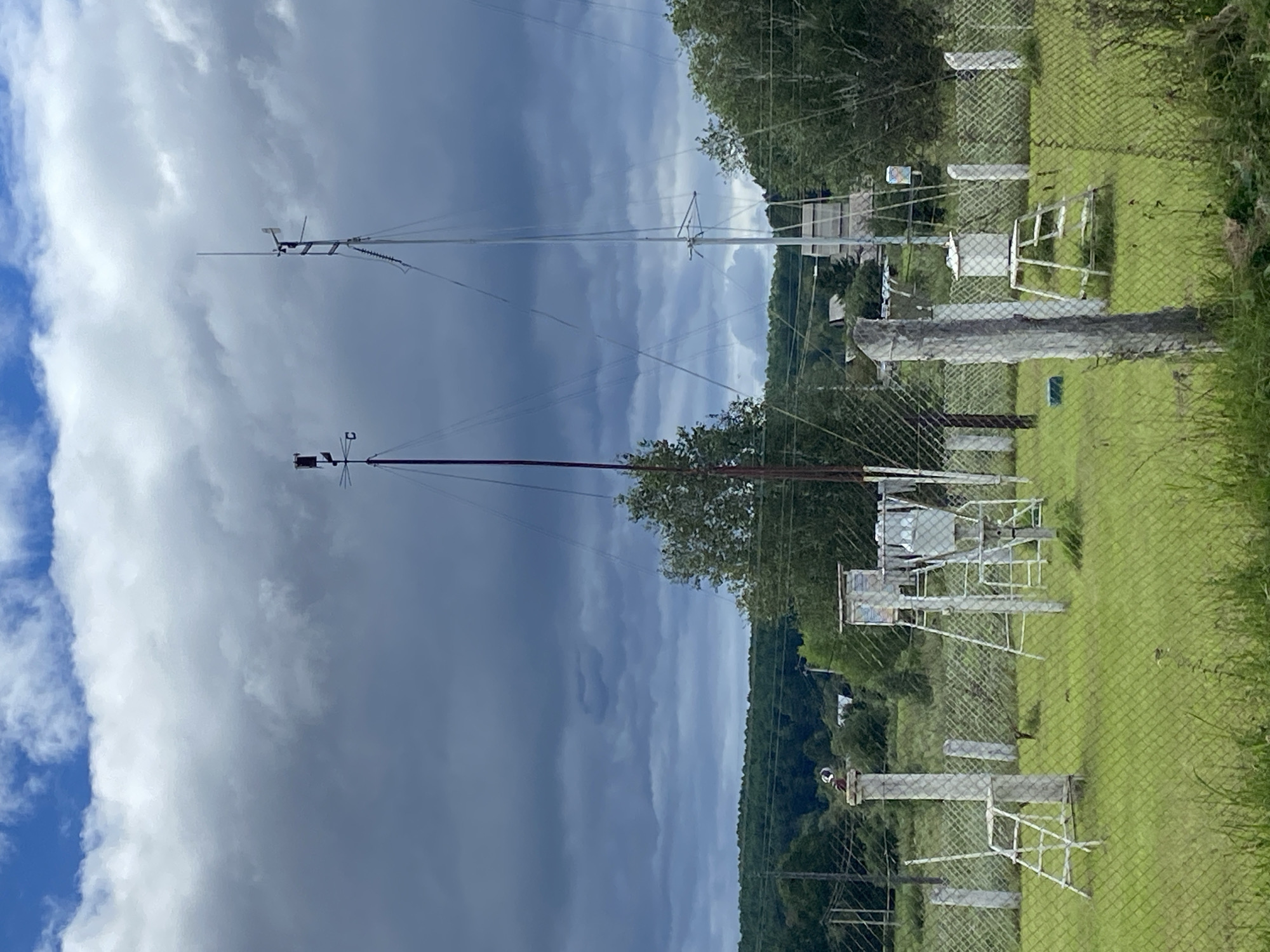 This image shows a meteorological station in Chernyayevo, Russia, under a cloudy sky. In the foreground, a chain-link fence encloses a grassy area containing various weather monitoring instruments. These include several white, ladder-like structures, some topped with small enclosures, and other white pillars. Two tall masts, one with a weather vane and anemometer, dominate the background. Behind the station, a line of trees and a few buildings are visible, set against a backdrop of a dense forest and rolling hills. The sky is filled with dark, ominous clouds, suggesting impending rain or stormy weather.