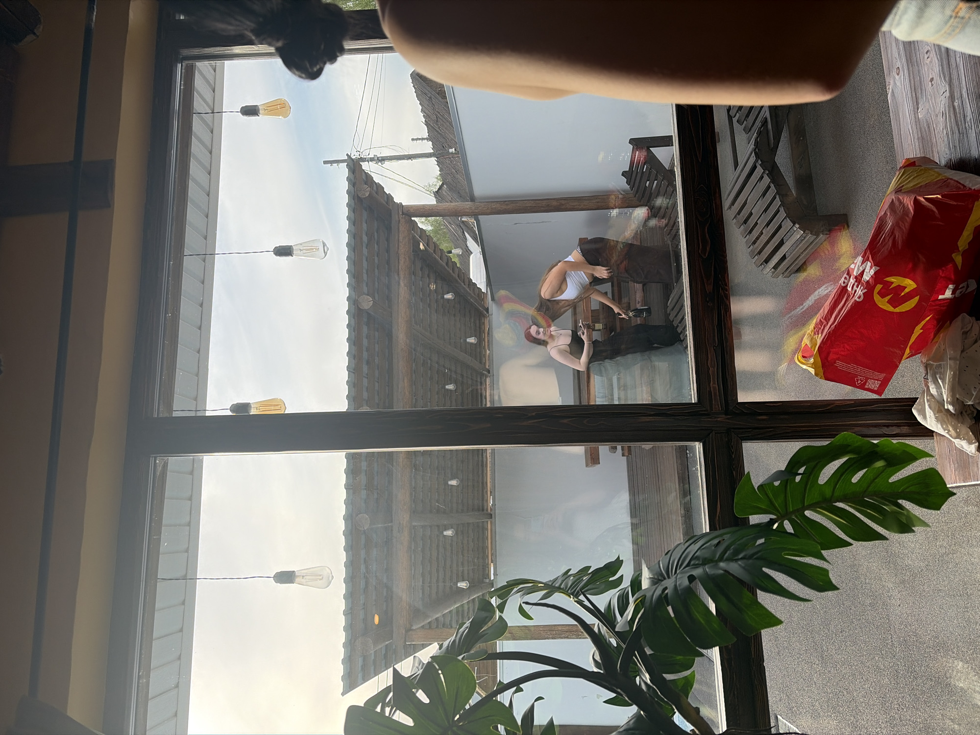 From an interior viewpoint in Старое Михайловское, Russia, the image captured through a window reveals two females in an exterior setting. One, dressed in a white top, is bending over while seemingly handing an object to the other, who's in a dark dress. The exterior setting also features overhead string lights along a rustic roof structure. The indoor environment includes a potted plant.