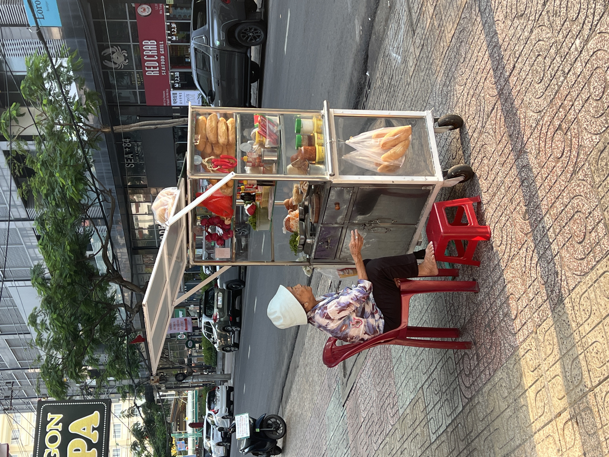 An elderly Vietnamese woman, wearing a floral print shirt, dark pants, and a white sun hat, sits on a red plastic chair next to a food cart on a tiled sidewalk. Her feet are bare, and her left hand is raised towards the cart. The food cart displays various items including baguettes, condiments, and what appears to be meat products. Behind the cart, a street is visible with parked cars and motorbikes. Buildings line the street, including establishments named "SEA +84," "RED CRAB Seafood Grill," and "Loro Boutique." The location is the city of Nha Trang, Vietnam. A sign with the words "GON PA" in large yellow letters is visible on the lower left.