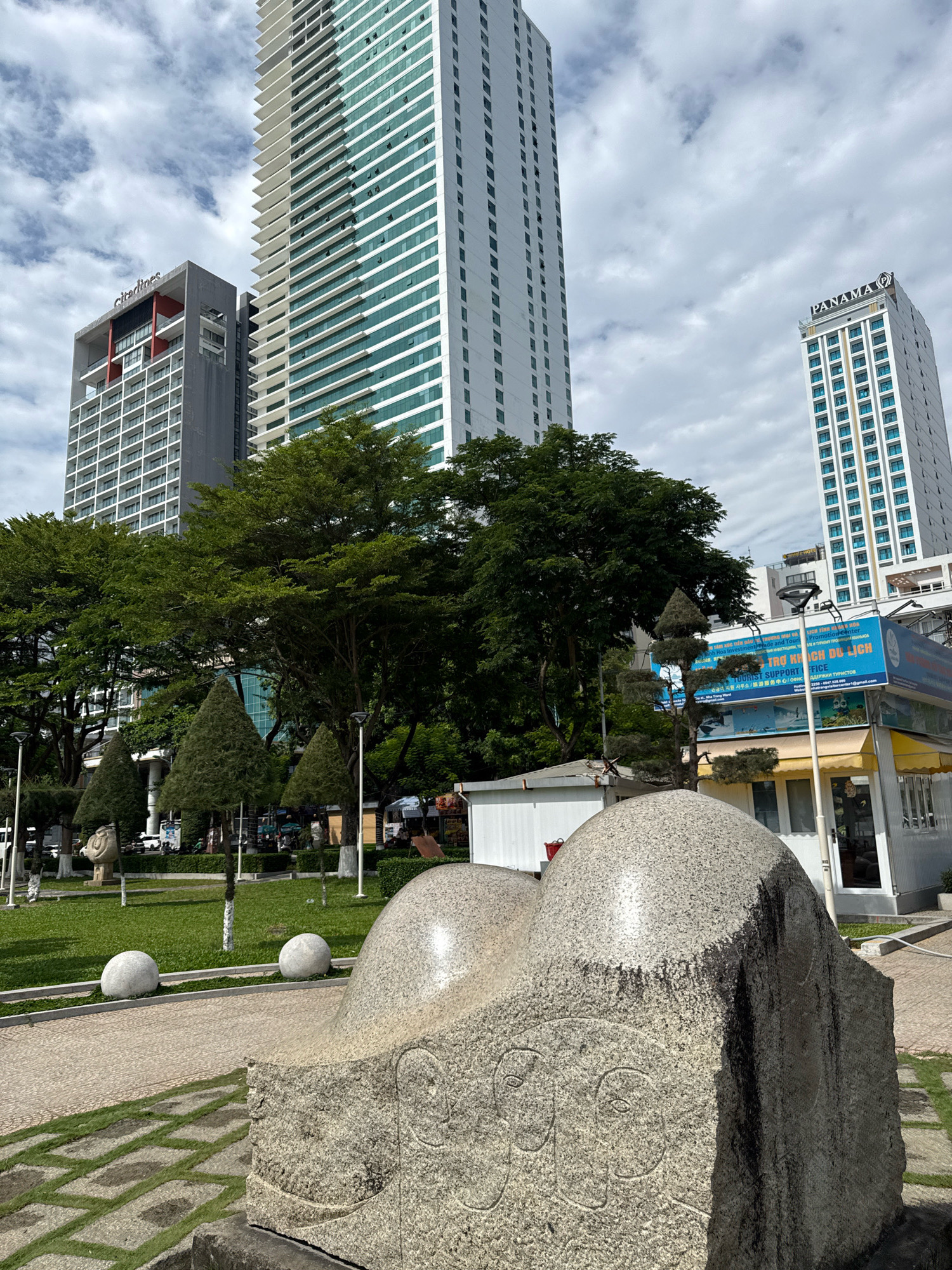 The image depicts a public space in Nha Trang, Vietnam, featuring a prominent stone sculpture in the foreground. The sculpture is a textured, grayish boulder with carved faces and abstract shapes. Behind it, a neatly manicured green lawn is dotted with spherical white planters and minimalist lampposts. In the mid-ground, several conical evergreen trees are artistically pruned and arranged. Beyond the trees, a vibrant blue and yellow building, possibly a tourist information center or shop, is visible. Towering in the background are modern high-rise buildings. To the left, a grey and white high-rise with the "Citadines" logo is identifiable. To the right, another tall white building with a blue-tinted facade bears the "PANAMA" logo. The sky is partially cloudy, suggesting daytime conditions. The scene appears to be a well-maintained urban park or plaza, characteristic of tourist areas in a coastal city.