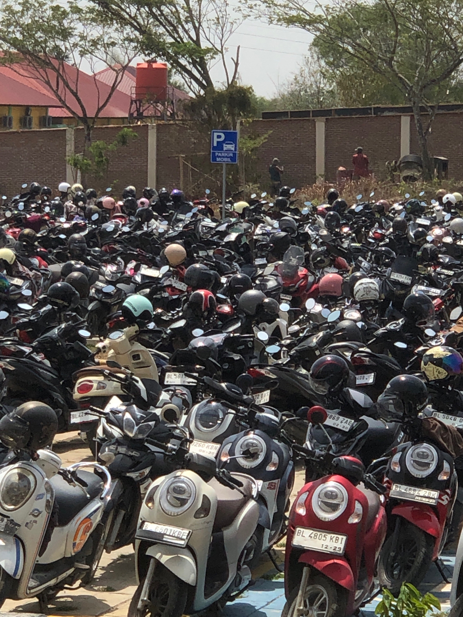 In Reuleut Timu, Indonesia, a densely packed outdoor parking lot is filled with a multitude of parked motorcycles. The scene is dominated by the numerous scooters, predominantly in dark colors like black and gray, with some splashes of red and white. Many of the motorcycles have helmets placed on their seats or handlebars. In the background, a brick wall runs horizontally across the frame, topped with a line of trees and a building with a red roof. A blue parking sign with a car icon is visible on a pole. Two individuals are standing near the brick wall, one wearing a dark shirt and the other a red garment. The ground appears to be paved, likely asphalt or concrete, and the overall lighting suggests a sunny day. The sheer volume of motorcycles indicates a busy area, possibly near a commercial or public facility.