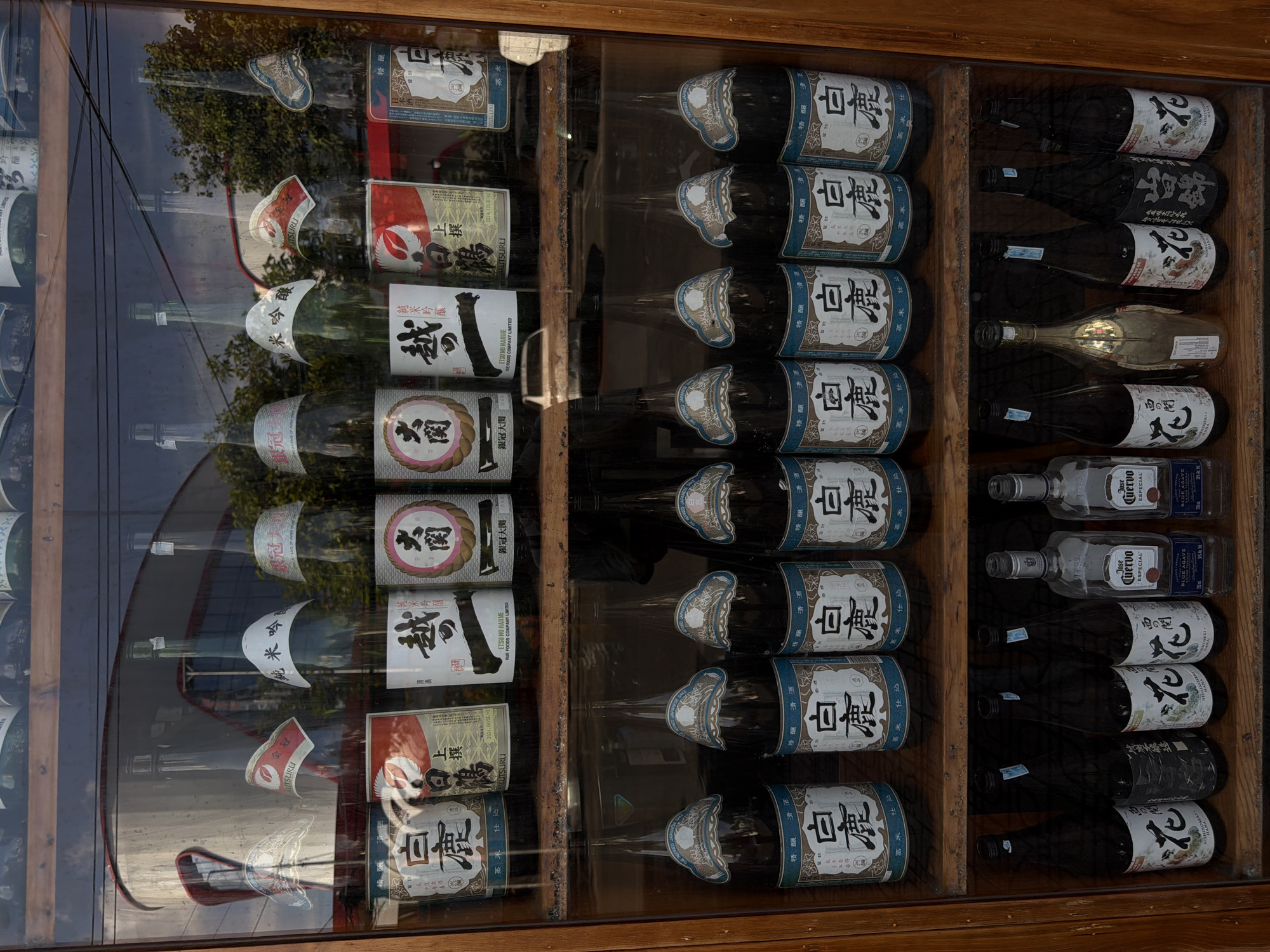 The image shows a display cabinet of alcoholic beverages, likely in a retail setting. The cabinet is made of wood and has glass doors. The shelves are filled with bottles of sake and tequila, mostly Japanese rice wine called "Sake". The bottles are arranged in neat rows and columns. The labels are visible and written in both Japanese and English. The environment is a reflection of the street, with greenery and buildings visible in the glass. The location might be a store in Nha Trang Ward, Vietnam. The scene is static, with no visible actions or interactions. The lighting is bright and even.