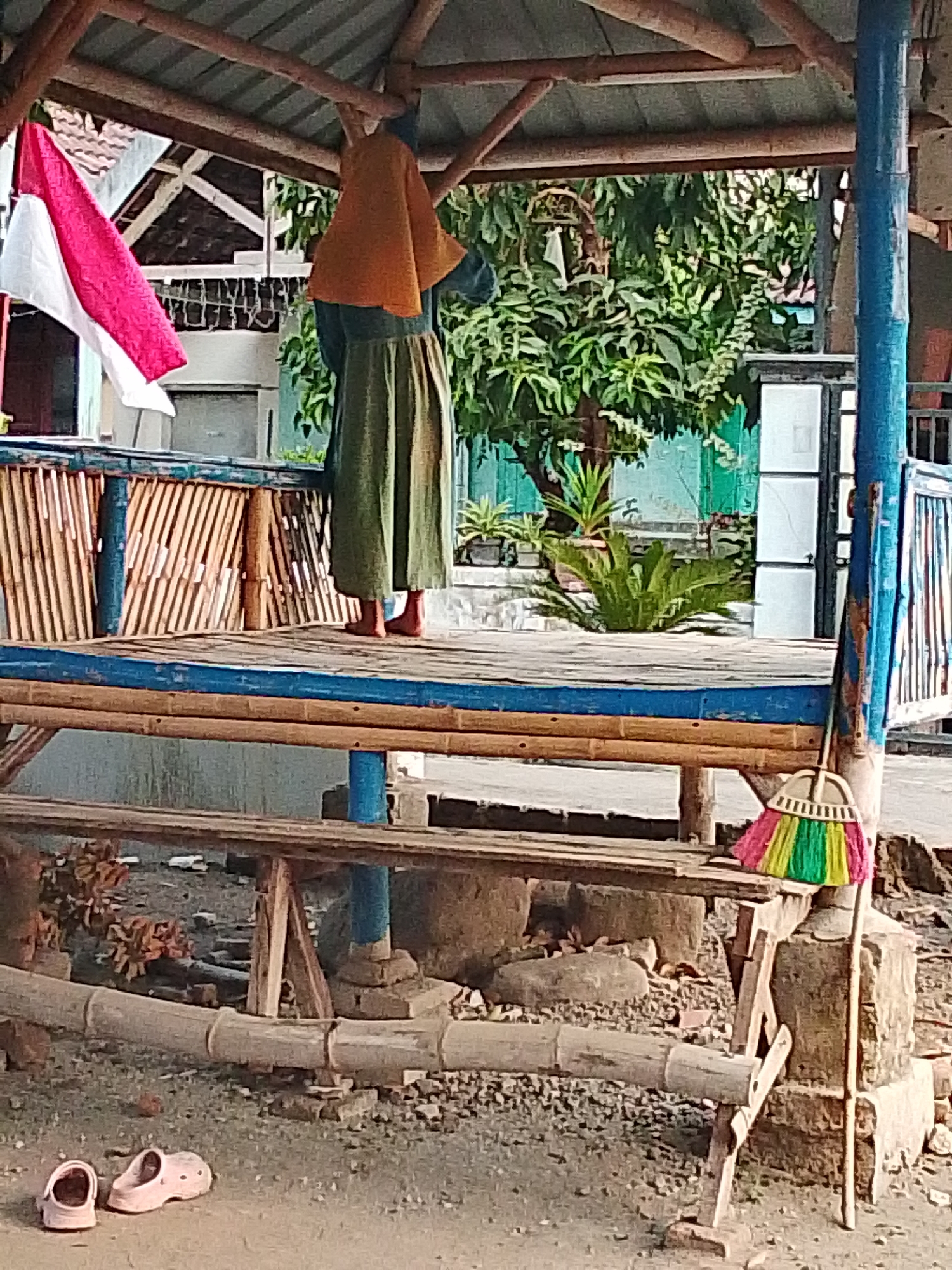 An outdoor scene depicts a person standing on a raised bamboo platform in what appears to be a shaded, open-air structure. The person, facing away from the camera, is wearing an orange headscarf and a long, green skirt. Visible on the platform is a flag, predominantly red and white, and a broom with multi-colored bristles leaning against a blue-painted post. Below the platform, on the ground, a pair of pink sandals are visible. The ground is covered with gravel and dirt, with visible bamboo structures supporting the platform. In the background, lush green trees and a building with a white facade are present. The overall impression is of a simple, rustic outdoor area, possibly in a village setting in Bulurejo, Indonesia, given the national flag and common building materials.