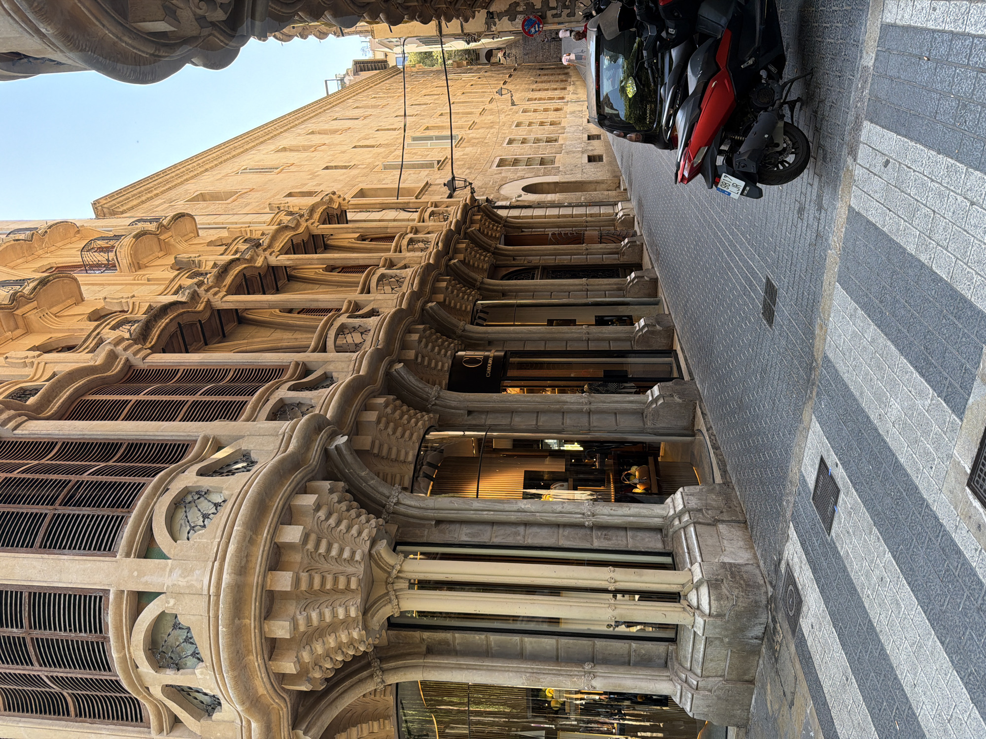 The image depicts a street scene in Palma, Spain, with a focus on the intricate architecture of a building on the left. The building features elaborate stone carvings, arched windows with louvred shutters, and decorative columns supporting a projecting facade. The street is paved with cobblestones and features a pedestrian crossing with alternating grey and white stripes. Two motorcycles, one red and black and another black, are parked on the right side of the street. A black car is also parked further down the street. The street narrows as it recedes into the distance, with more buildings visible on the right. The sky is clear and blue. Several people are visible walking up the street in the distance. The overall impression is of a charming, historic European streetscape.