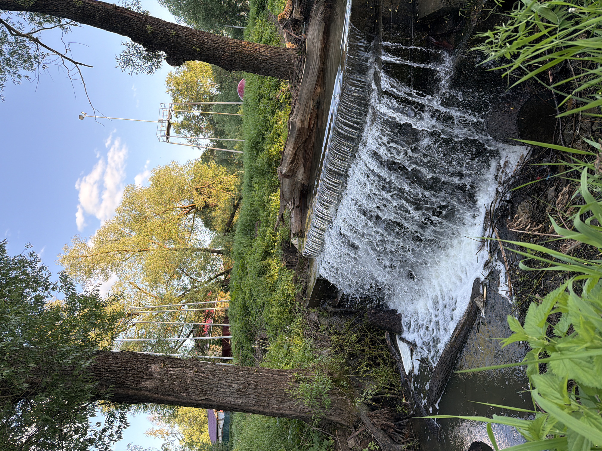 The image shows a small waterfall in a park-like setting. The water cascades over a ledge constructed of logs and concrete, creating a textured, frothy flow. The immediate area surrounding the waterfall is overgrown with greenery, including grasses and other plants. Trees frame the scene on both sides. In the background, a structure with white metal supports is visible against a backdrop of foliage and a blue sky. The photo appears to be taken in Sin'kovo, Russia.