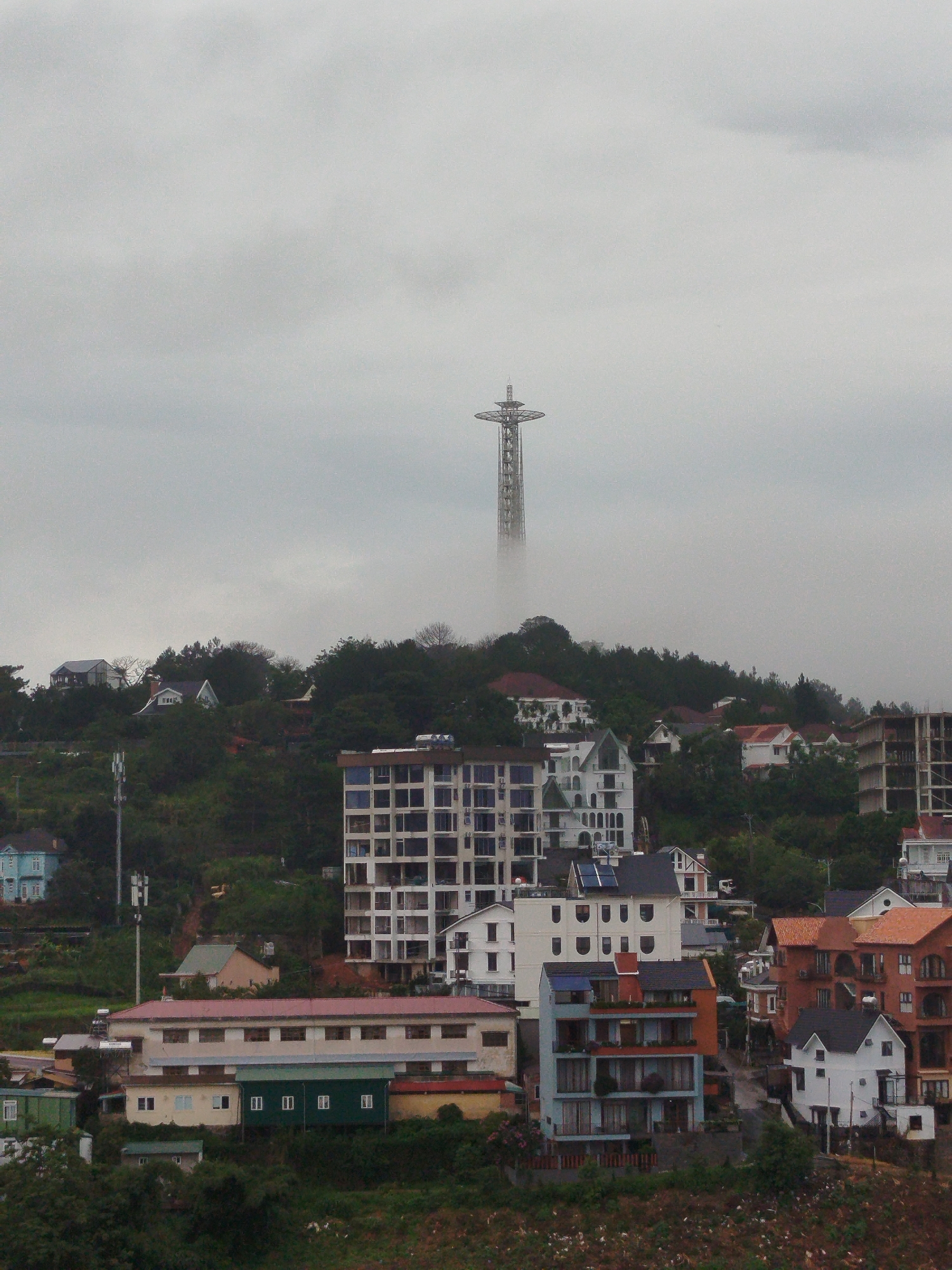 The image depicts a panoramic view of a hillside city, with a distinctive tower structure prominently visible in the distance, partially obscured by fog. The scene is characterized by a dense cluster of buildings, varying in architectural style and size, interspersed with lush green vegetation. In the foreground, several multi-story buildings, some appearing modern with glass facades and others with more traditional designs, are visible. The hillside terrain is evident, with buildings built on different levels. The sky is overcast and hazy, suggesting a cloudy or misty day. The tower in the background appears to be a telecommunications tower or a monument, its metallic structure intricate and towering over the surrounding landscape. Based on the distinctive tower and the hilly terrain covered with buildings, this scene is identifiable as being in Da Lat, Vietnam, likely near the Linh An Pagoda, which features a large statue of Quan Am and is situated on a hill.