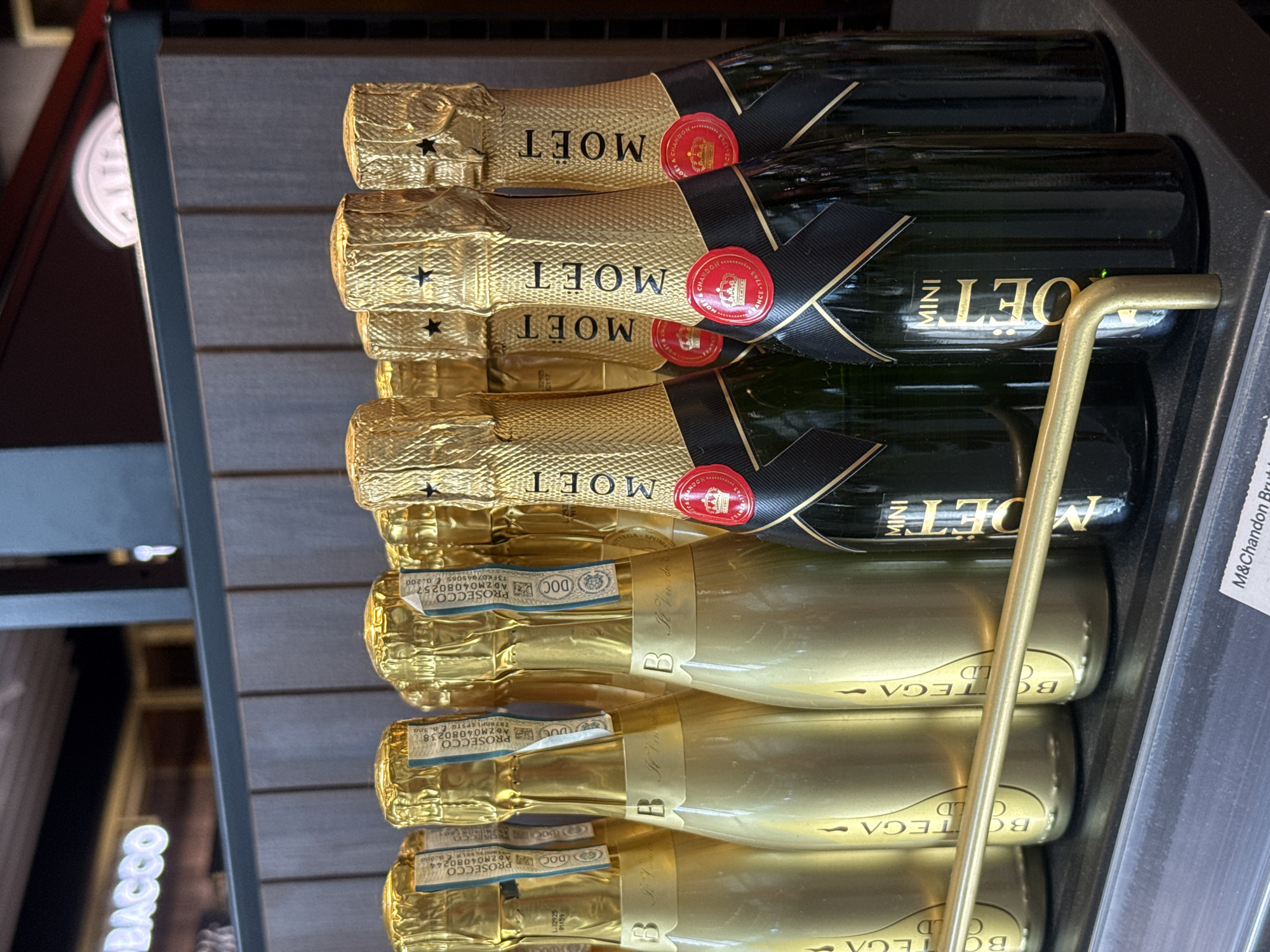 A close-up, eye-level shot showcases a shelf filled with miniature bottles of sparkling wine. The bottles are arranged in two distinct groups. On the left, a row of gold-foiled bottles, likely Bottega Gold, with "B" embossed on the neck and "Il Vino dei Poeti" script are visible. Some have "PROSECCO DOC" labels. On the right, several dark green bottles, identified as Moët & Chandon Mini Champagne, are positioned. These bottles feature black labels with gold lettering spelling "MOET" vertically and a red circular seal with a crown. A thin, golden metal bar runs horizontally across the foreground, partially obscuring the lower portion of the bottles. The background consists of a textured wooden wall and a blurred illuminated sign that reads "BACCO". The image suggests a retail environment, possibly a wine shop or a duty-free store in Sofia, Bulgaria.