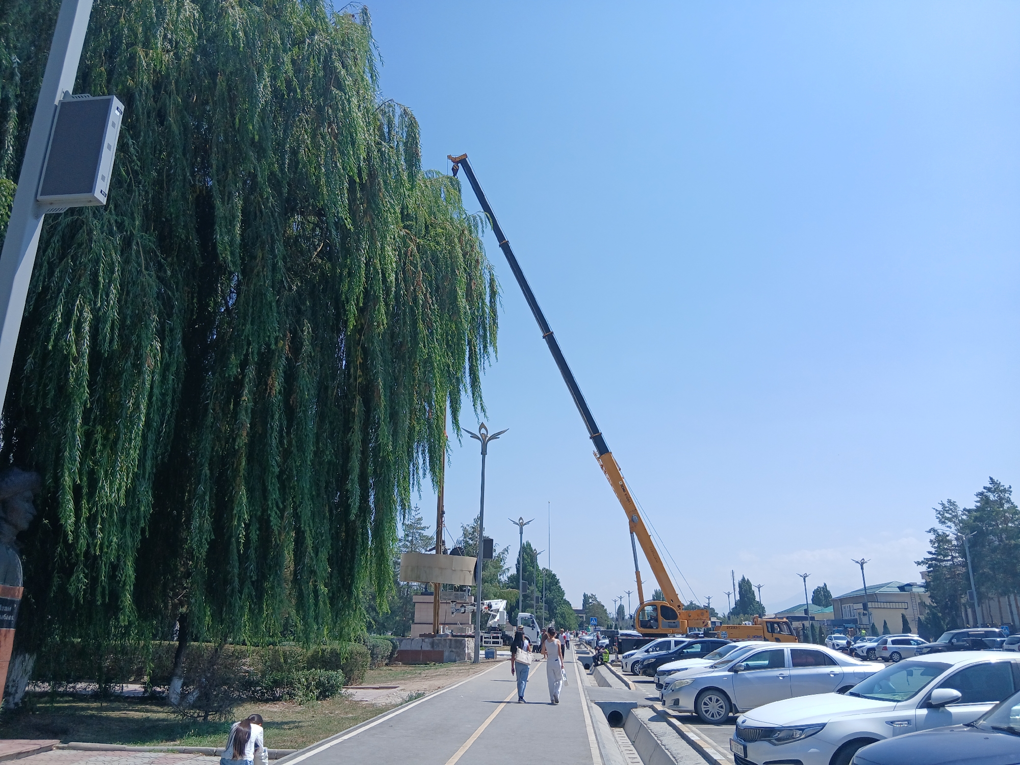 In Talas City, Kyrgyzstan, on a clear day, a long, dark crane arm extends from the right side of the frame, reaching high into the bright blue sky, angled over a row of parked cars and a pathway. The crane appears to be yellow at its base. To the left, a large weeping willow tree dominates the foreground, its dense green foliage cascading down, partially obscuring a grey pole with a small rectangular dark grey device attached to it. A statue's base is visible to the far left, with some inscription on it. A paved pathway runs horizontally across the mid-ground, where two individuals, one in a white outfit and another in blue jeans and a dark top, are walking away from the camera. Further down the pathway, more people are visible, as well as a white truck and other construction equipment. The pathway is bordered on the right by a parking lot filled with numerous cars of various colors, including white, silver, and dark grey. In the background, trees and buildings are present, with some mountains visible in the far distance under the expansive sky.