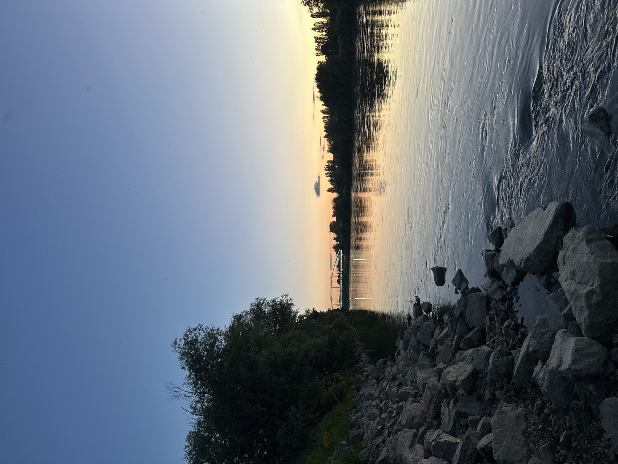 Here is a description of the image:
The photograph depicts a waterscape at dusk. A river with a slightly rippled surface reflects the warm hues of the setting sun. The shoreline is rocky, transitioning into a vegetated area with trees and bushes. In the background, a bridge is visible, its lights illuminating the water. The sky transitions from a clear blue to a gradient of orange and yellow near the horizon. The location is likely near the Sava River in the City of Zagreb, Croatia.