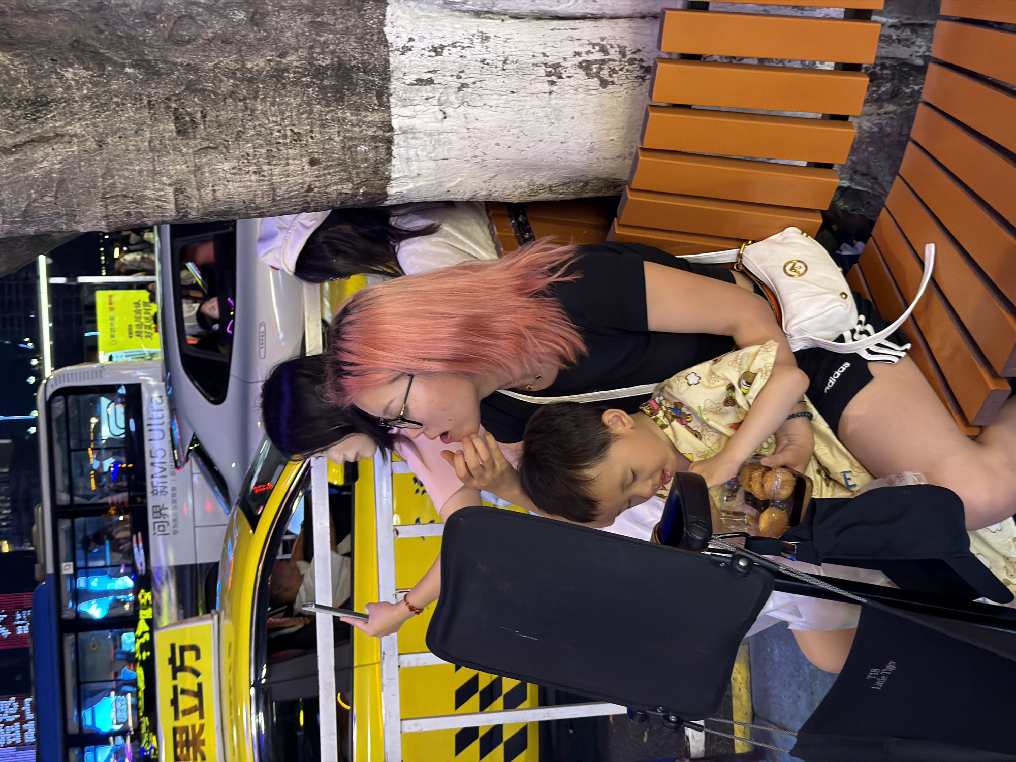 In the Yuzhong District of China, at street level at night, a young boy with dark hair sits on an orange slatted bench, holding a clear container with three brown pastry-like items. He is wearing a light-colored garment with a pattern of cartoonish animals. To his right, a woman with long, pink-dyed hair and glasses is looking down and to the left, her right hand near her mouth. She is wearing a black t-shirt and a white purse is slung across her body. Another woman, partially visible to the left, with long dark hair, is looking forward and holding a smartphone up, possibly taking a photo or video. Behind them, a yellow taxi is stopped, and further back, a silver car is in motion, with passengers visible inside. A bus is also visible in the background, with illuminated windows. A tree trunk with white paint on its lower section stands to the right of the frame. The scene suggests a busy urban environment at night, with traffic and people engaged in everyday activities.